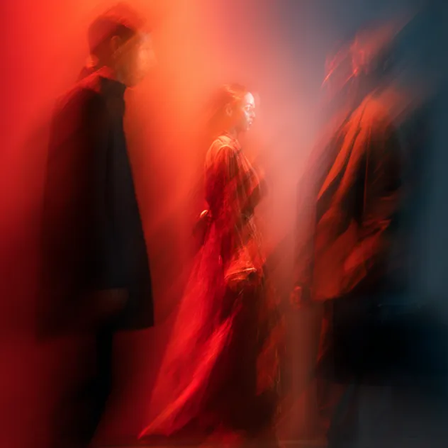Three people in motion with blurred effects, one in the center wearing a long red dress under red and dark lighting.
