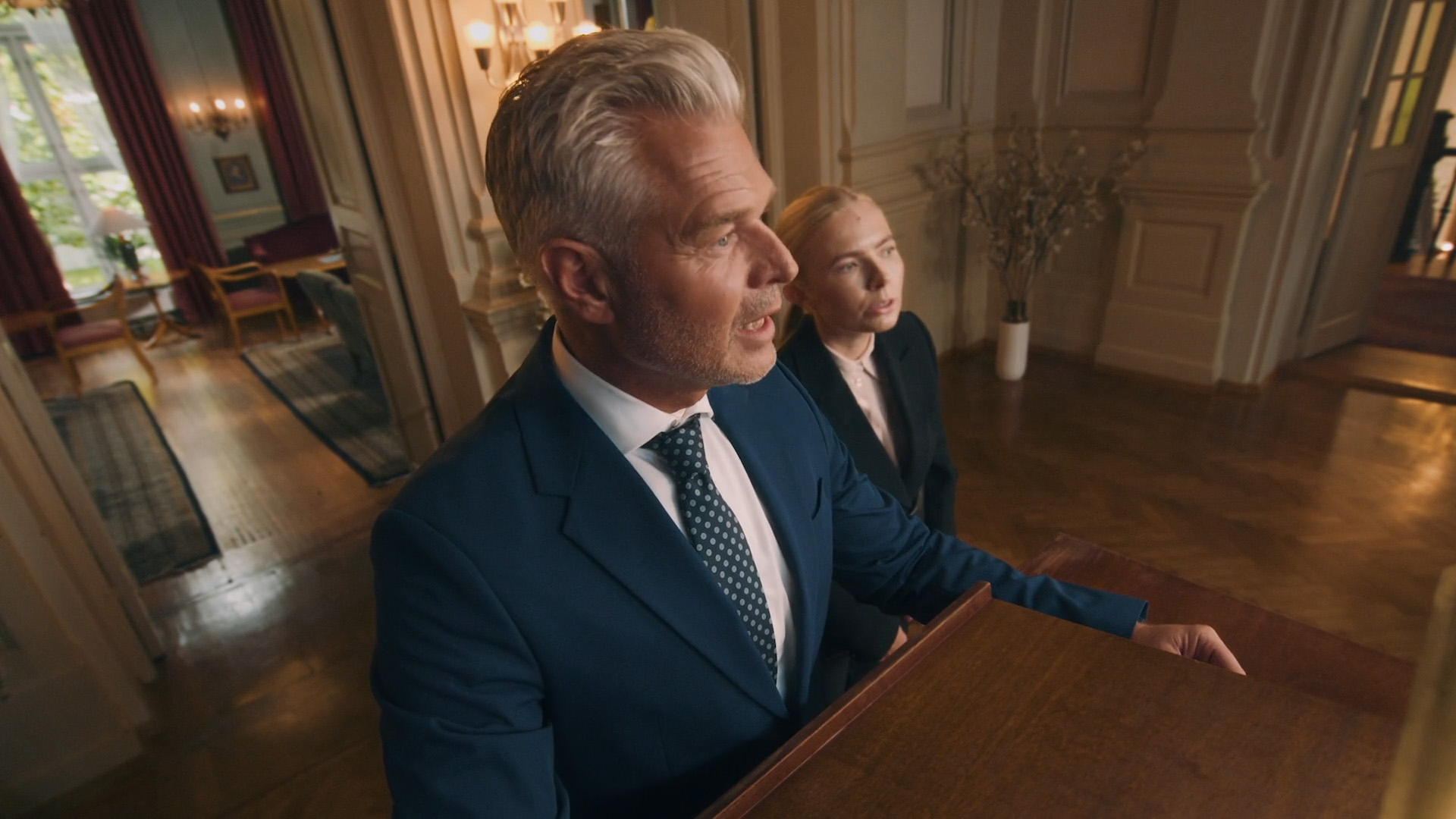 En mann og en kvinne i mørke dresser står ved en talerstol i et staselig rom.