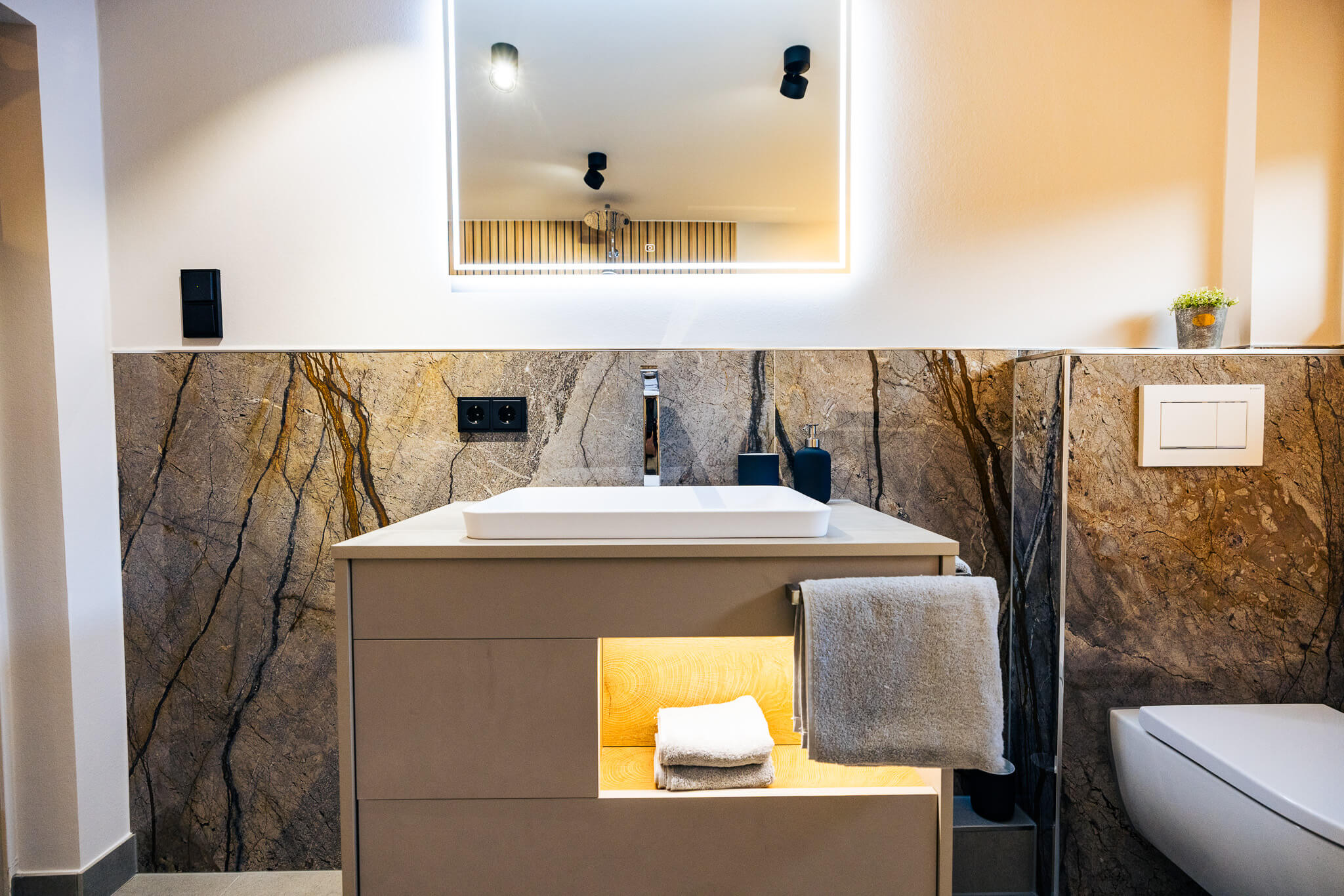 Moderner Badezimmerschrank mit beleuchtetem Fach, weißen Handtüchern, Waschbecken, Spiegel mit Licht und braun gemusterten Wandfliesen.