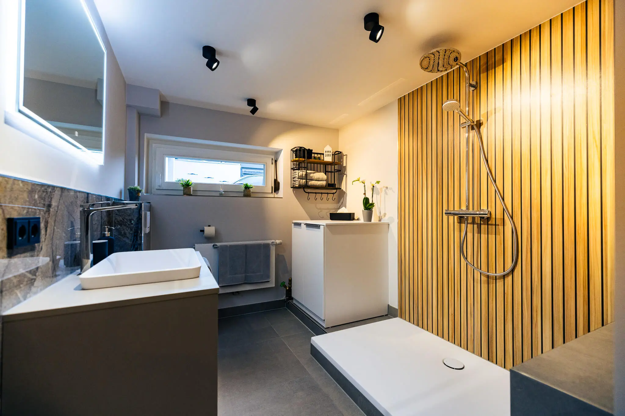 Modernes Badezimmer mit einer freistehenden goldfarbenen Badewanne, einer begehbaren Dusche mit Marmorwand, rundem Fenster und Holzregal mit Handtüchern.