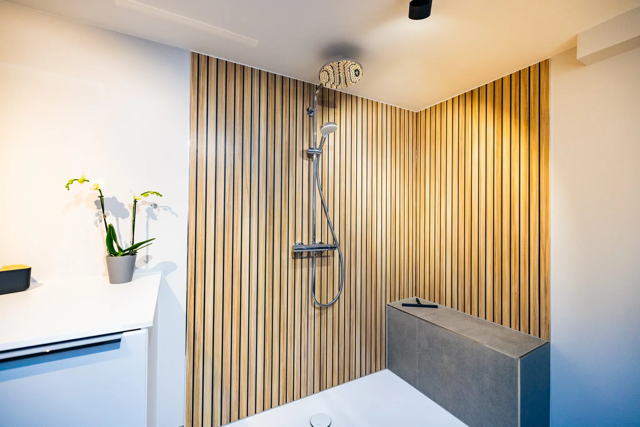 Barrierefreie Dusche mit modernem Regenduschkopf, Holzpaneelwand und grauer Sitzbank.