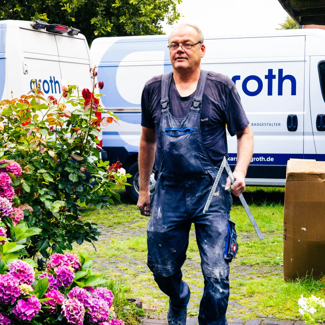 Ein Handwerker in schwarzer Arbeitskleidung läuft mit einem Winkelmesser in der Hand, im Hintergrund zwei Transporter mit der Aufschrift ‚groth‘ und blühende pinke und rote Blumen.