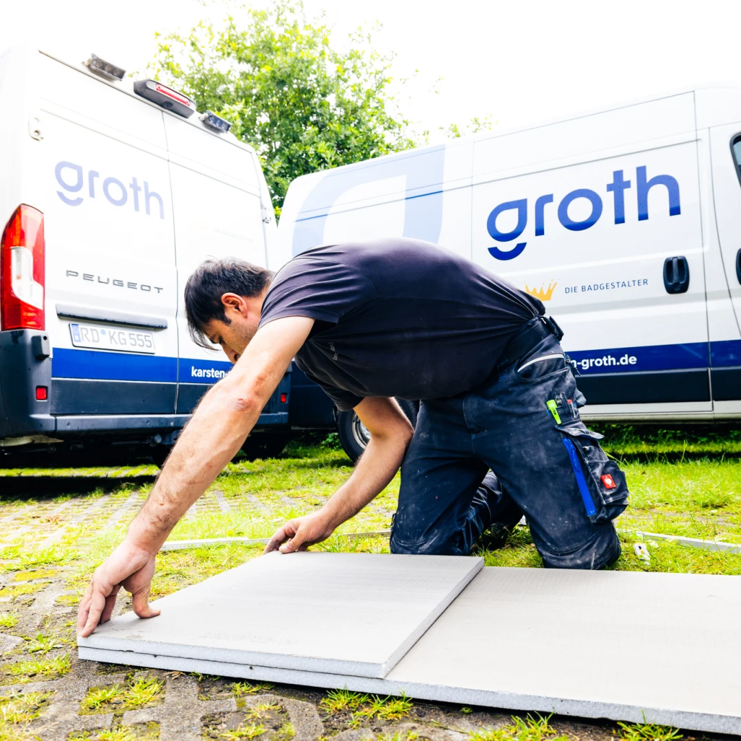 Handwerker kniet draußen und legt Fliesen neben zwei weißen Transportern mit Aufschrift Groth.