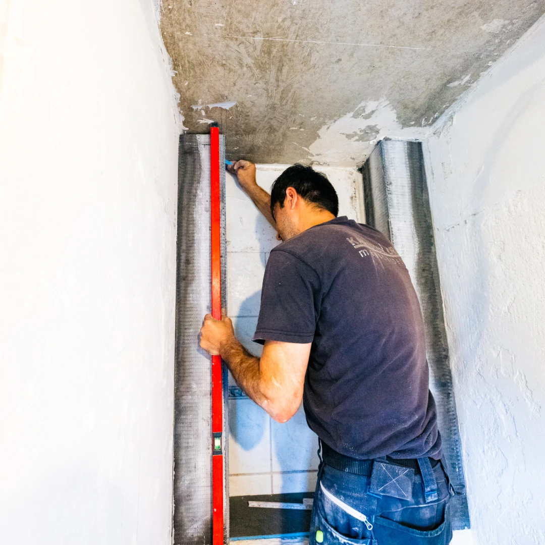 Handwerker misst mit einer Wasserwaage eine Wand in einem Raum mit weißem Putz und rohen Betonflächen.