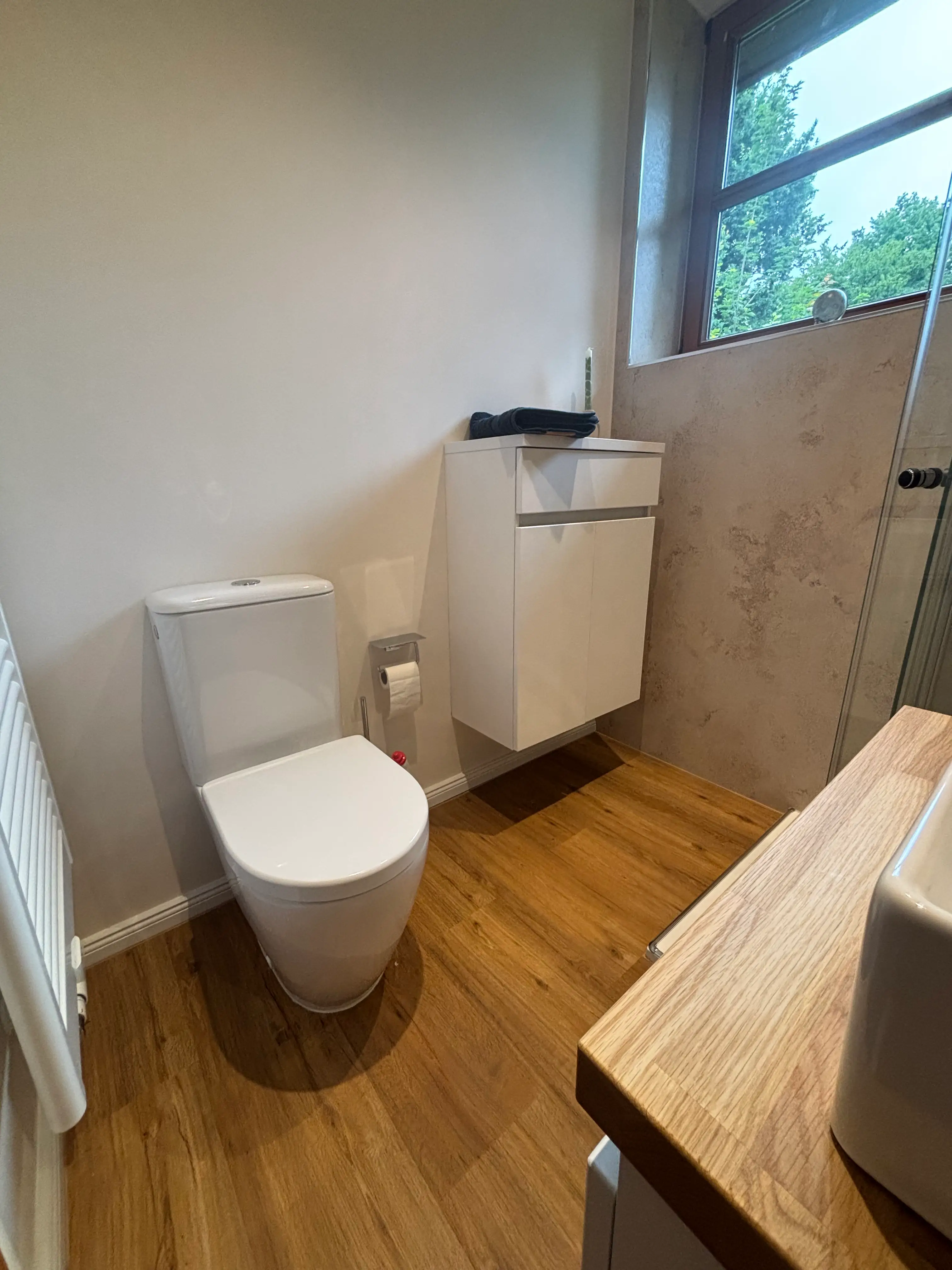 Kleines, modernes Badezimmer mit Holzfußboden, weißer Toilette, Wandwaschbecken und Schrank unter Fenster.