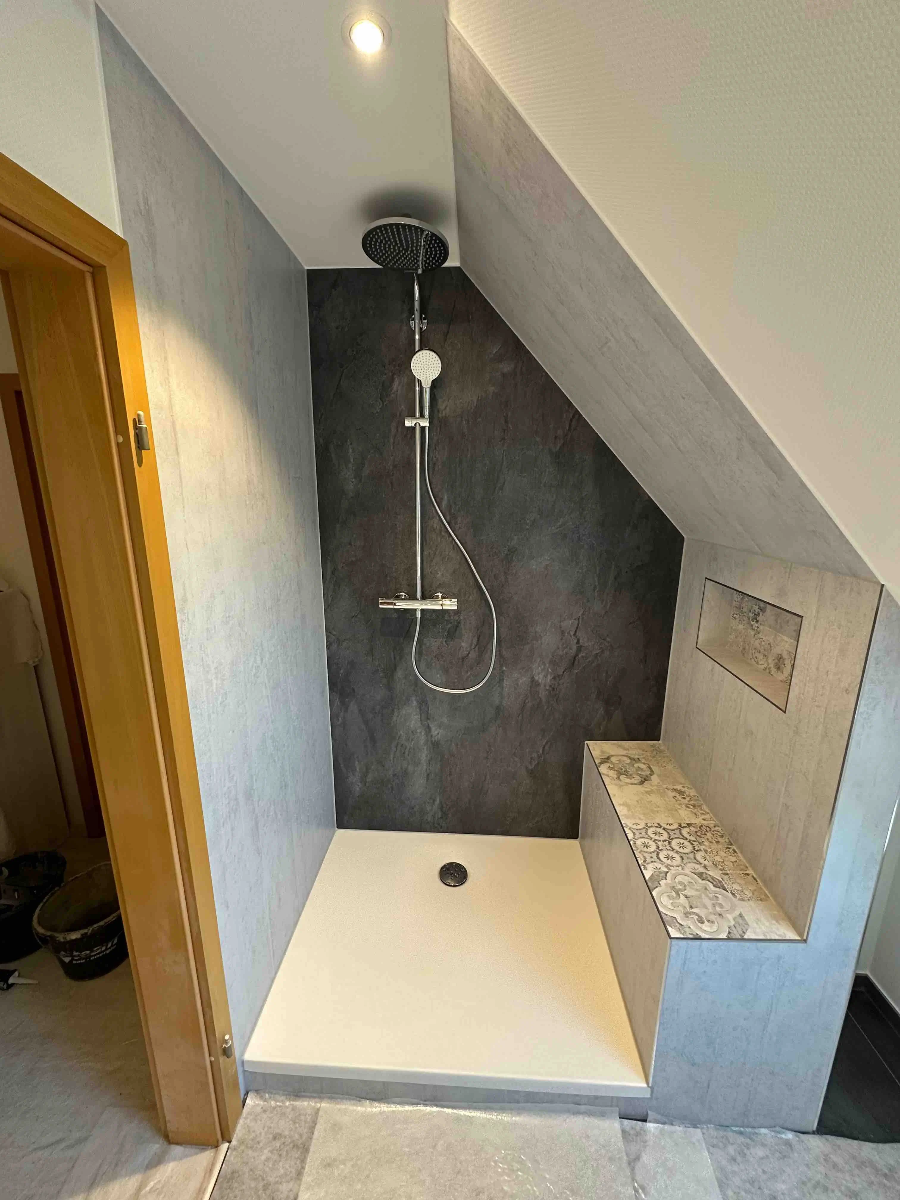 Dusche mit geneigter Decke, schwarzer Rückwand, großer Duschkopf, Handbrause und Sitzbank mit dekorativen Fliesen.