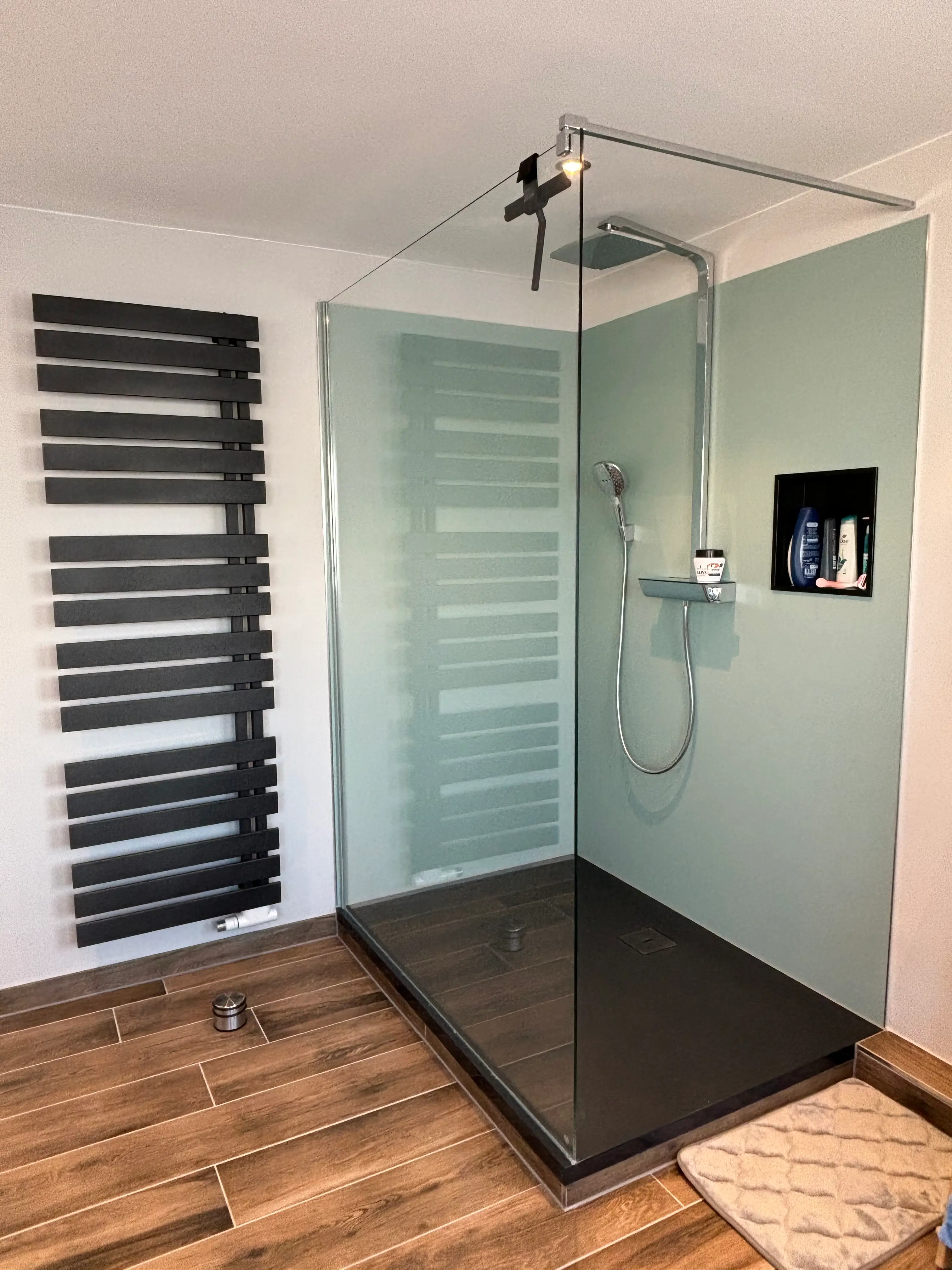 Moderne barrierefreie Dusche mit Glasabtrennung, Handbrause, integrierter Ablage und dunklem Wandheizkörper, Holzoptikfliesenboden.