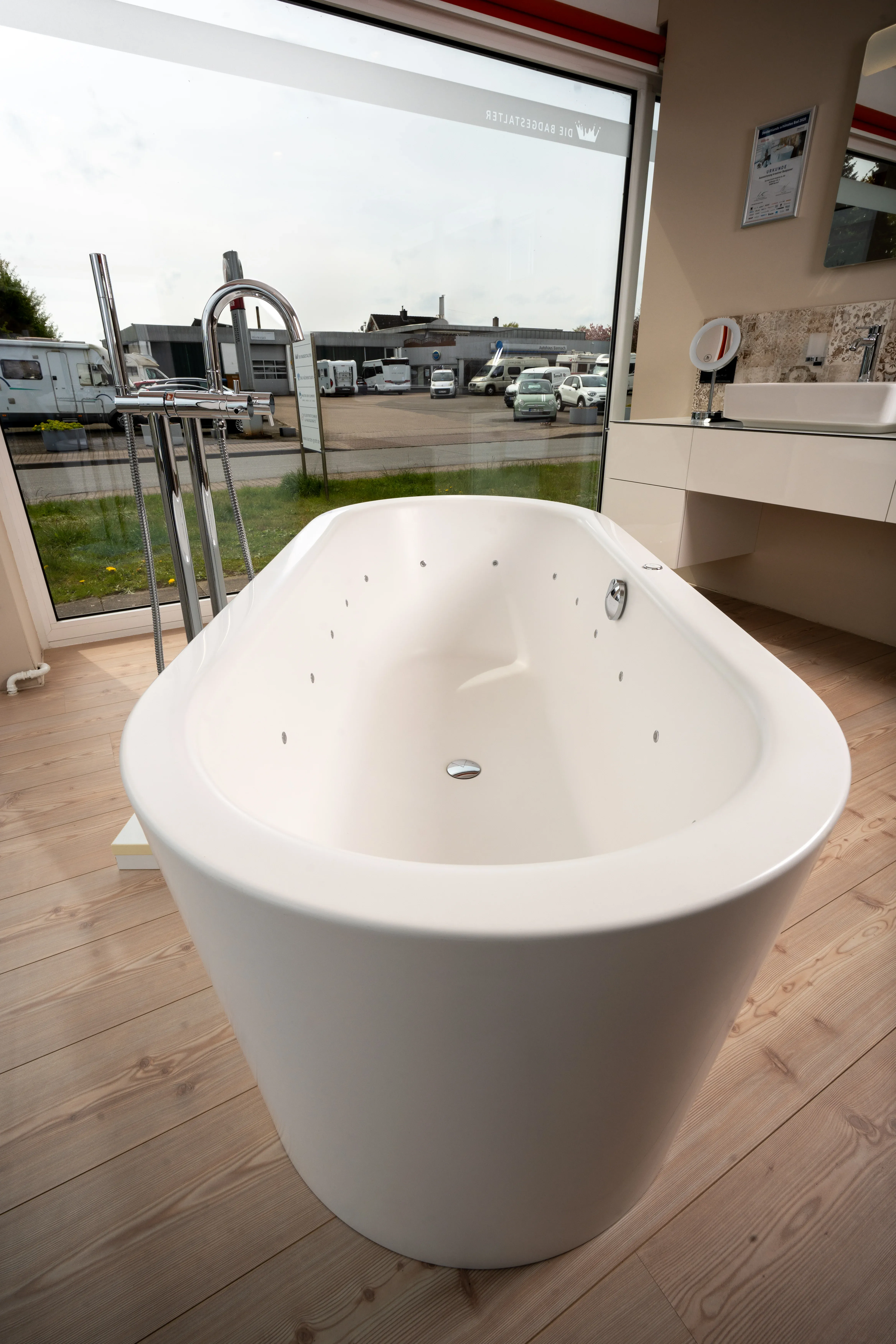 Freistehende weiße Badewanne mit Chrom-Wasserhahn vor großem Fenster mit Blick auf Parkplatz und Wohnmobile.