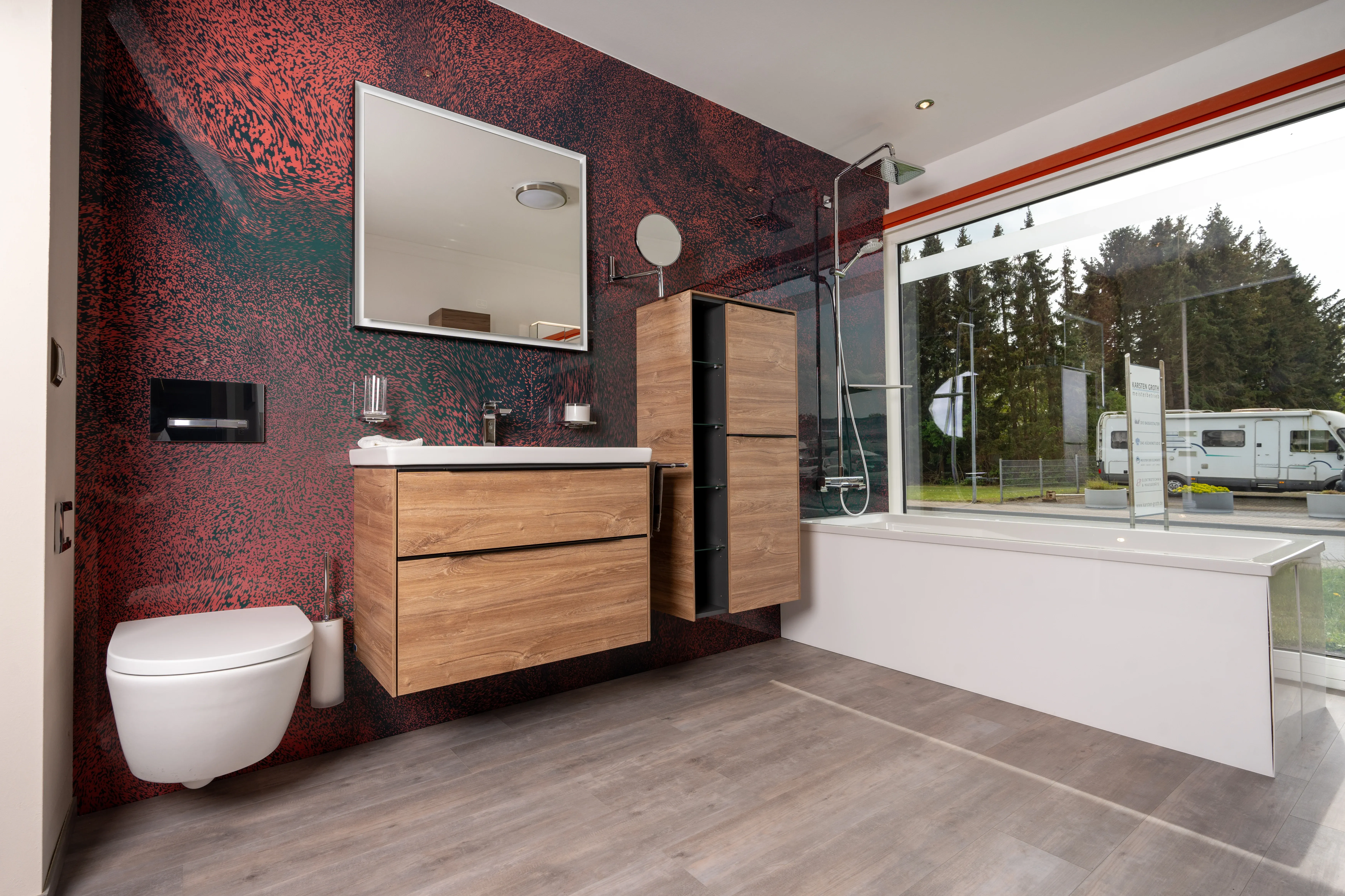 Modernes Badezimmer mit hängendem WC, Holzwaschtisch, Wandspiegel und Badewanne vor großem Fenster mit Blick auf Bäume und Wohnwagen.