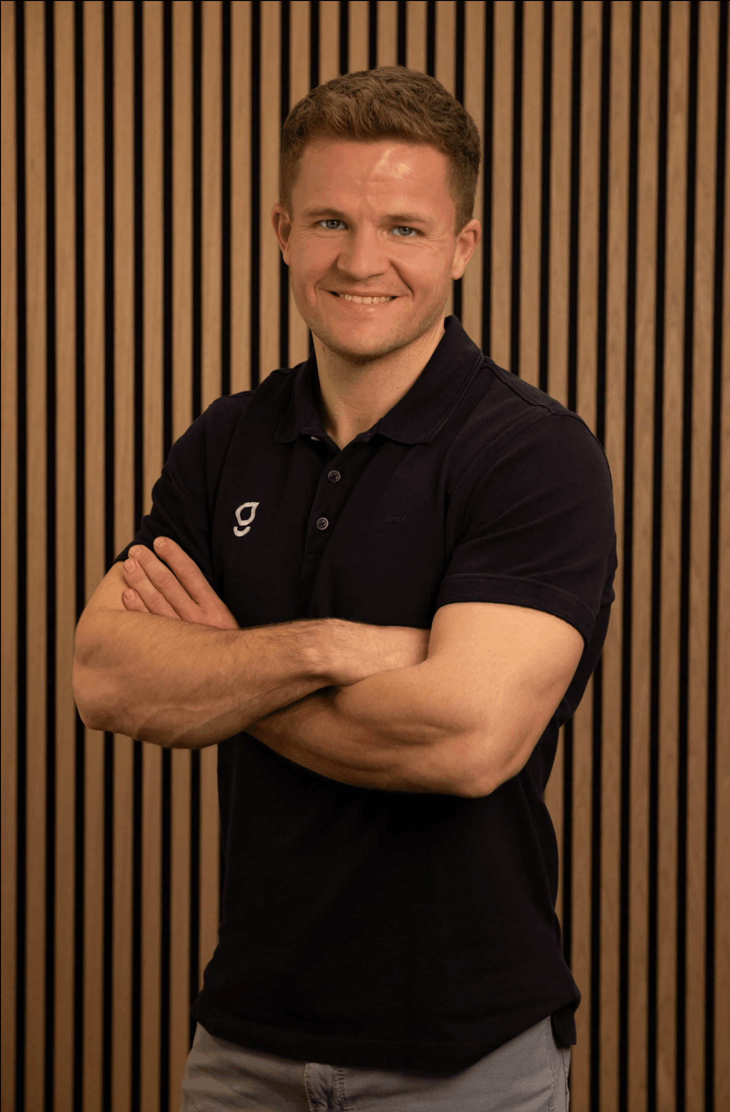Mann mit kurzen braunen Haaren und blauem Poloshirt steht vor einer Holzlattenwand mit verschränkten Armen und lächelt.
