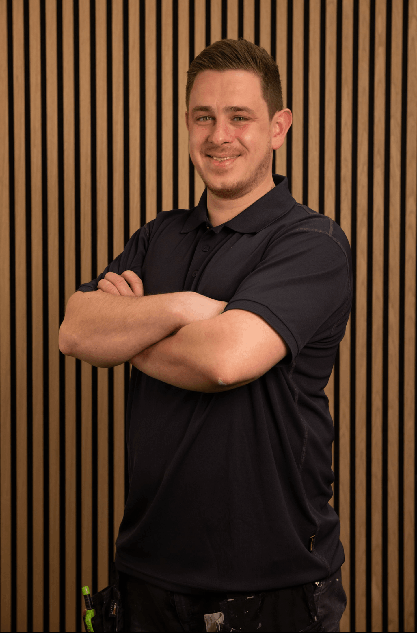 Junger Mann mit kurzem braunem Haar, der in einem schwarzen Polohemd vor einer Wand mit vertikalen Holzstreifen lächelt und die Arme verschränkt hat.
