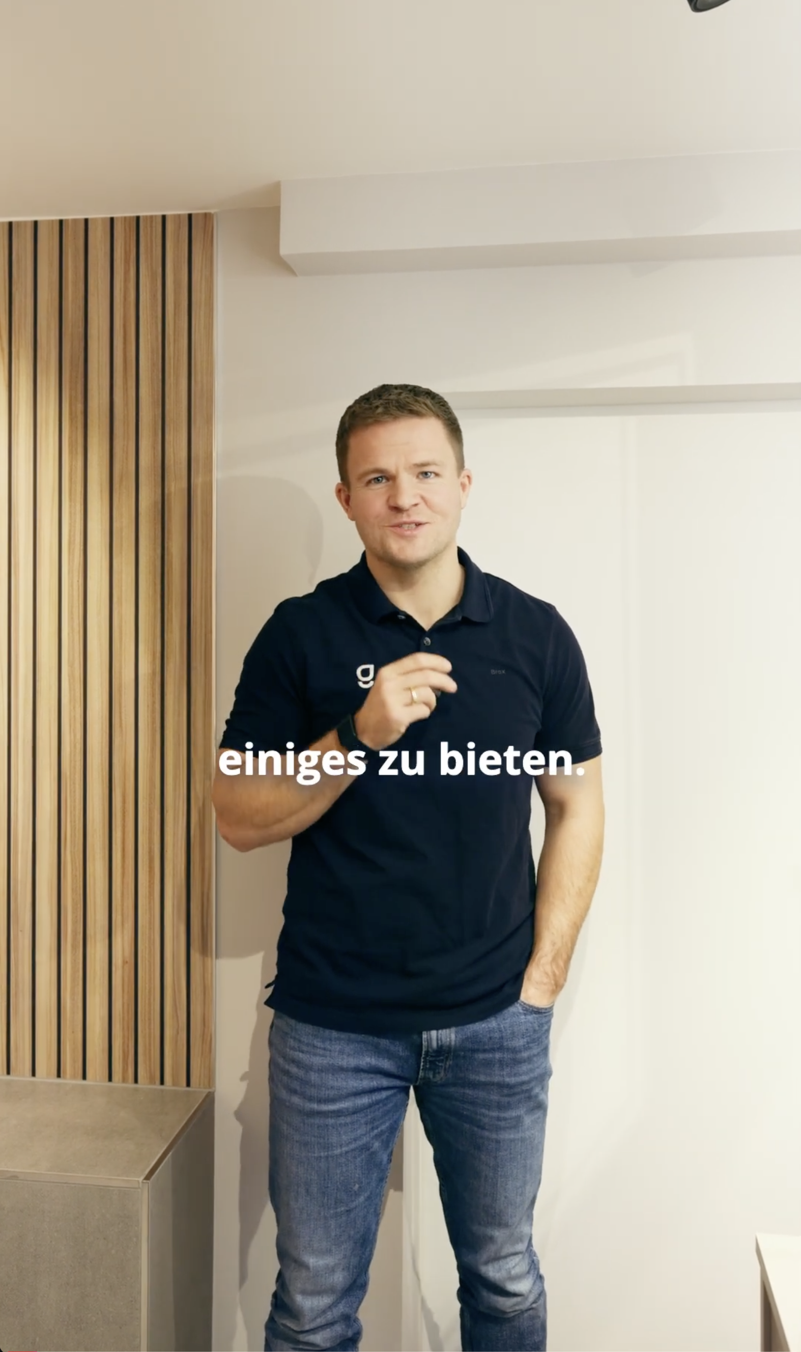 Lächelnder Mann mit blauer Poloshirt und Smartwatch steht in einer modernen Badezimmerumgebung mit Waschbecken und Holzverkleidung.