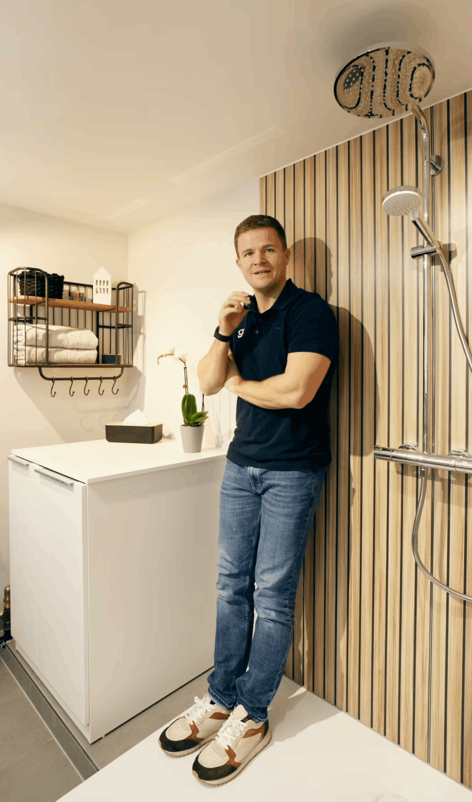 Lächelnder Mann mit blauer Poloshirt und Smartwatch steht in einer modernen Badezimmerumgebung mit Waschbecken und Holzverkleidung.