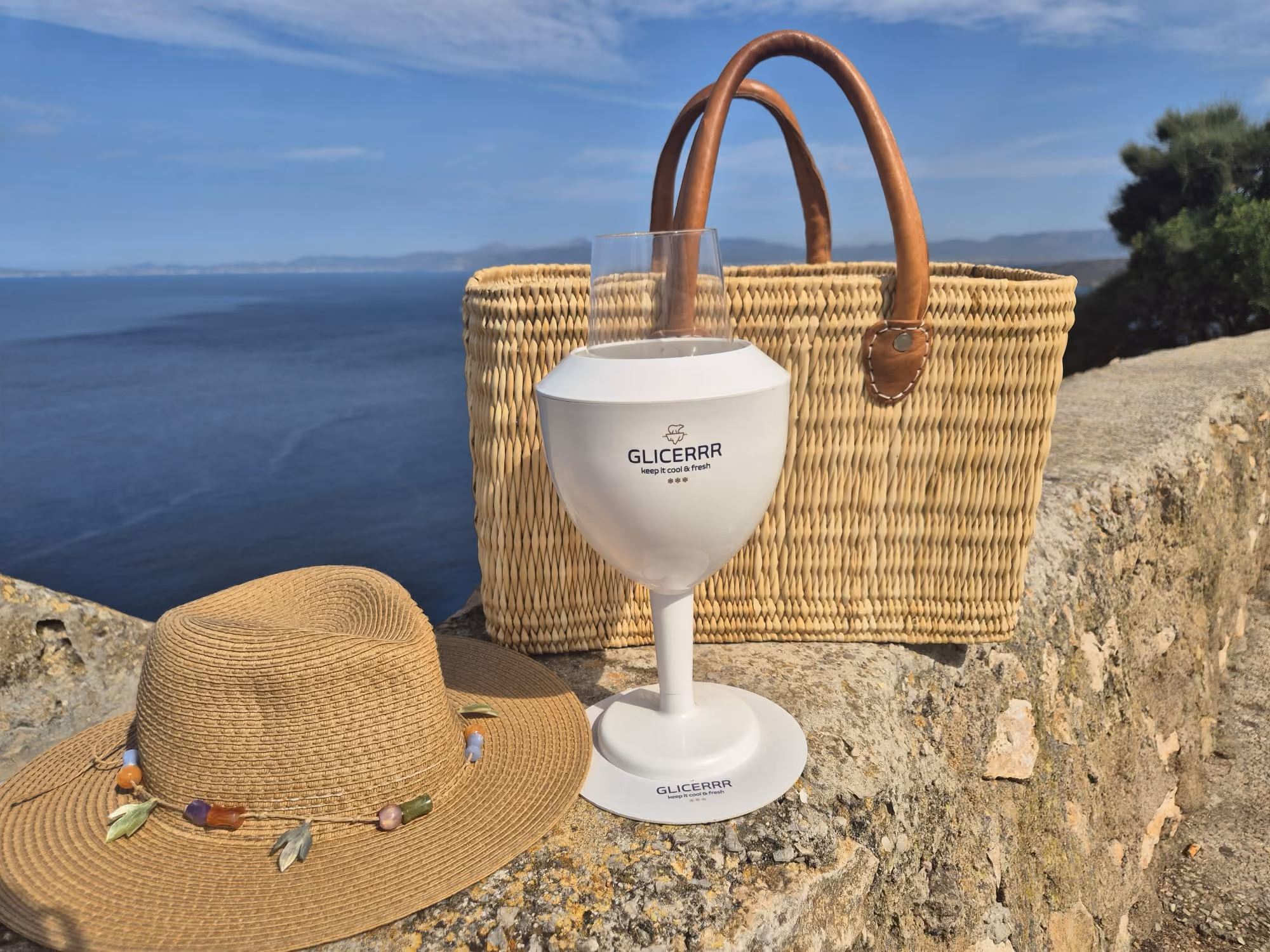 A white GLICERRR branded insulated wine glass cooler on a stone ledge next to a woven straw handbag with leather handles and a straw sunhat decorated with colorful beads, with a calm sea and partly cloudy sky in the background.