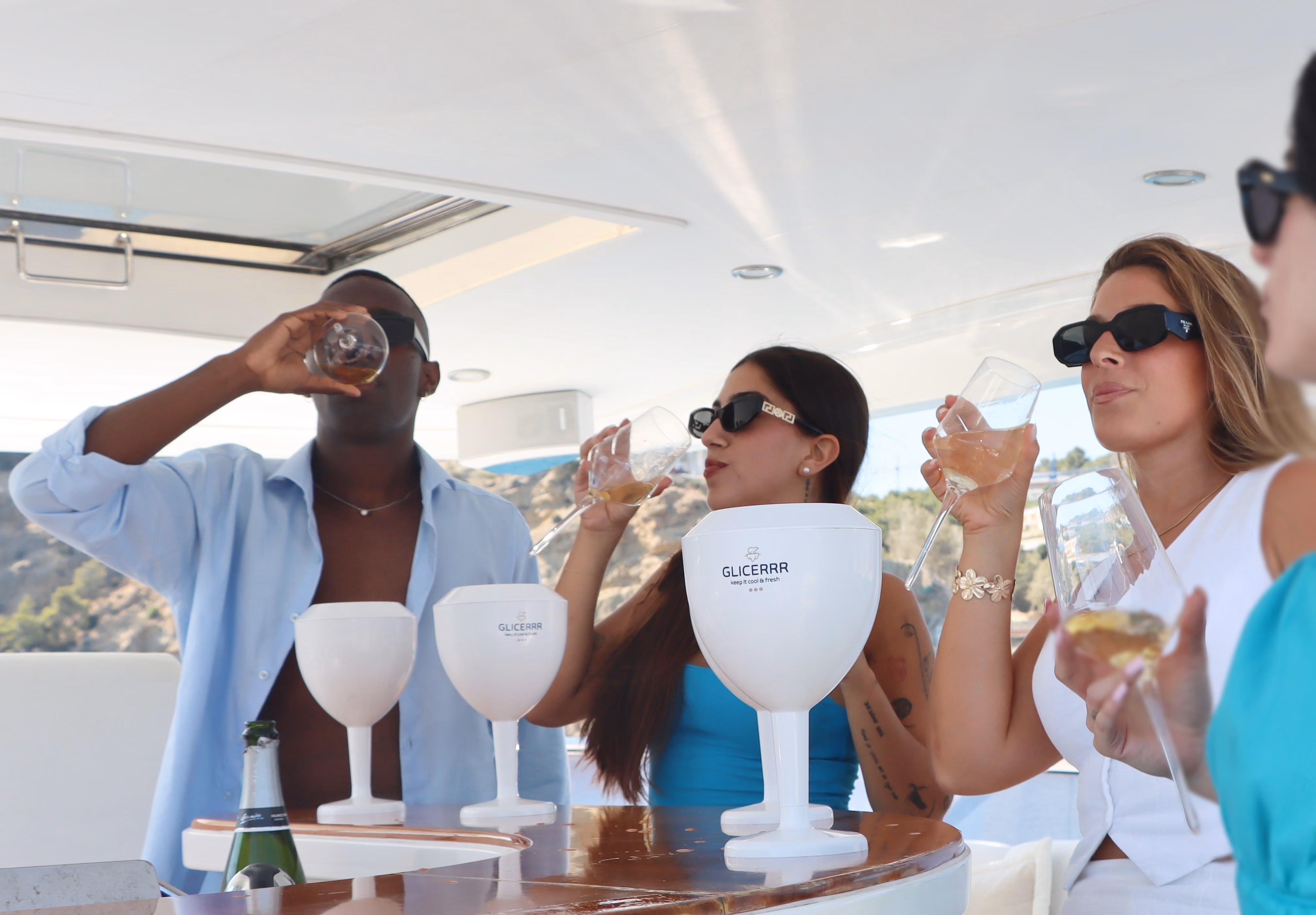 Four people wearing sunglasses enjoying drinks in a stylish setting, with white GLICERRR wine glass coolers on the table.