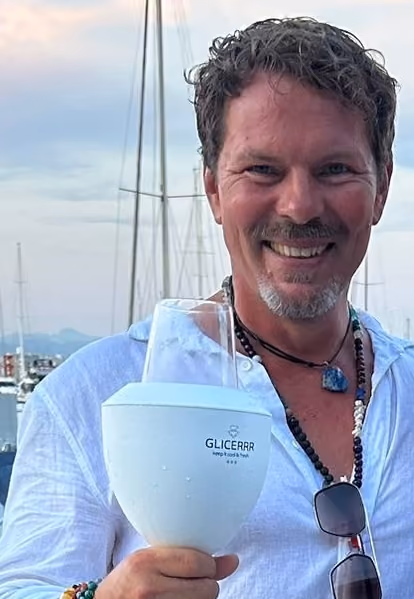 Smiling man with curly hair and a goatee wearing a white shirt and beaded necklace, holding a white GLICERR wine cooler with a glass inside, with a marina and boats in the background.