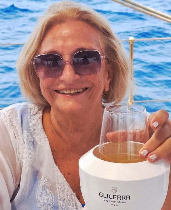 Smiling older woman with short blonde hair wearing large sunglasses and a white lace top, holding a glass with a light brown beverage placed in a white GLICERRR wine glass cooler, with a blue ocean background indicating she is on a boat.