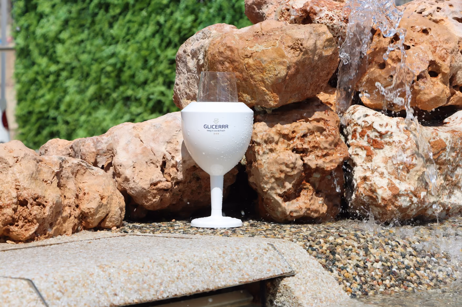 A white plastic wineglass cooler with the text 'GLICERRR' on it, placed on a stone surface near a rock waterfall with water flowing beside it.