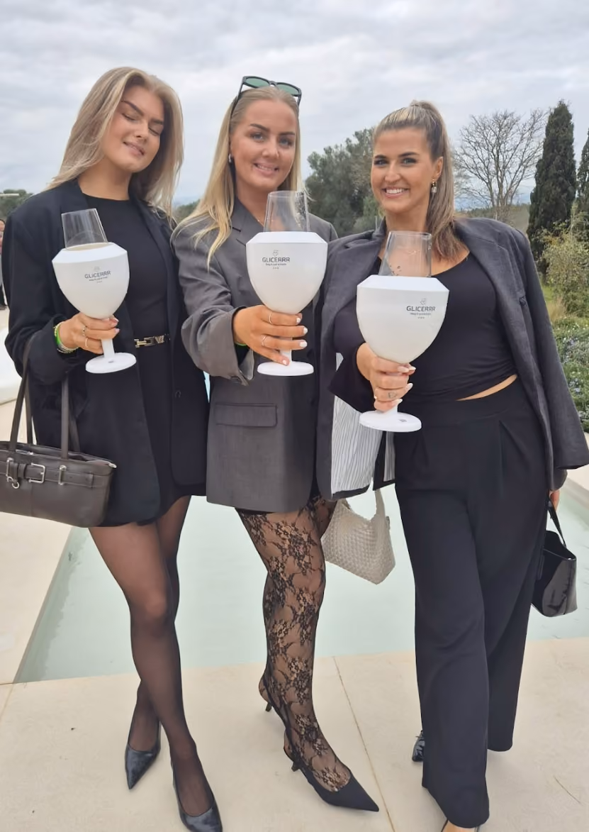 Three women standing outdoors near a pool holding a white wine glasses cooler on them, dressed in black and gray outfits, smiling at the camera.