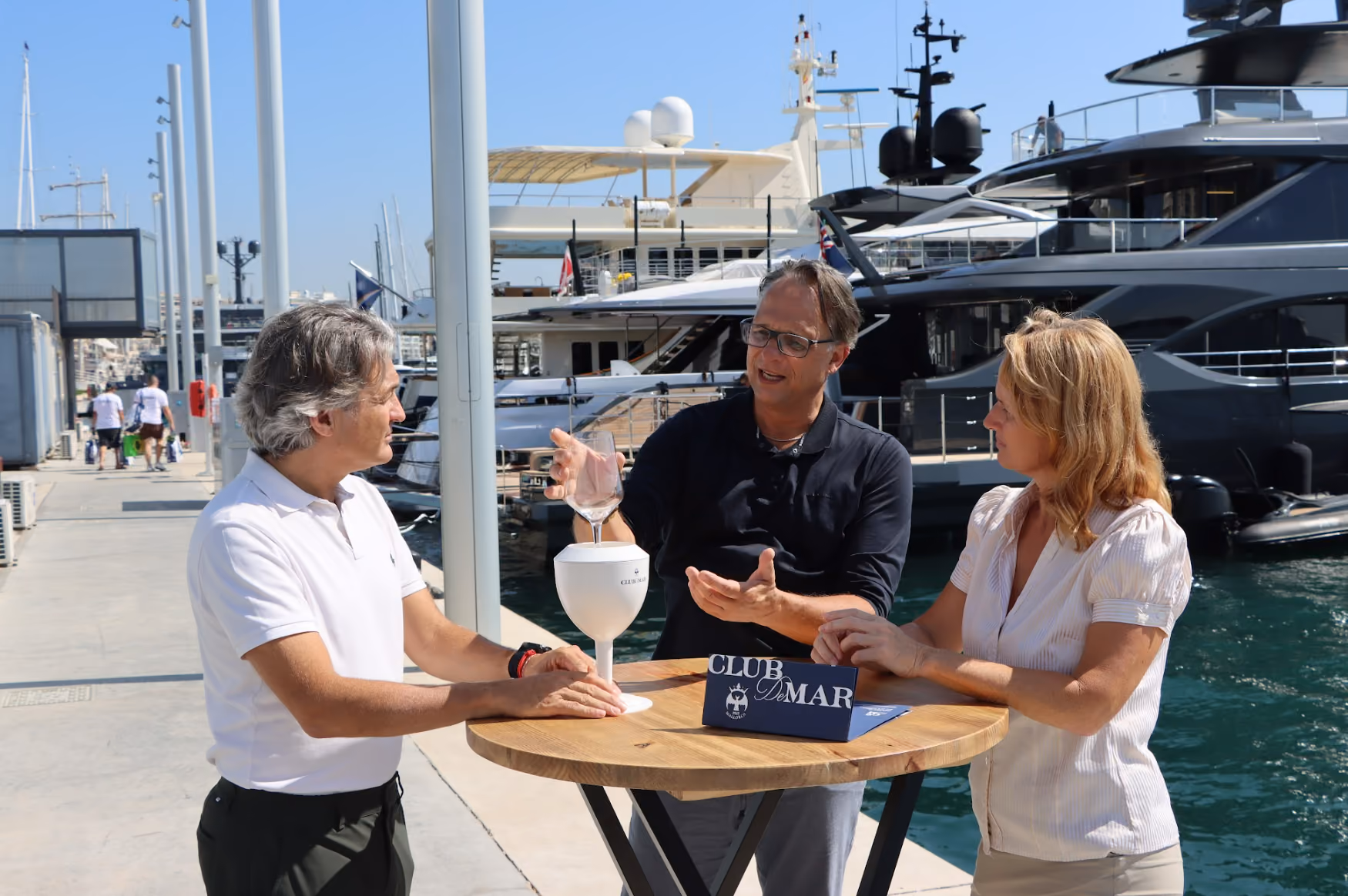 Three people are standing at a round wooden table by a marina, engaged in conversation under a clear blue sky, with large yachts in the background and a GLICERR on the table.