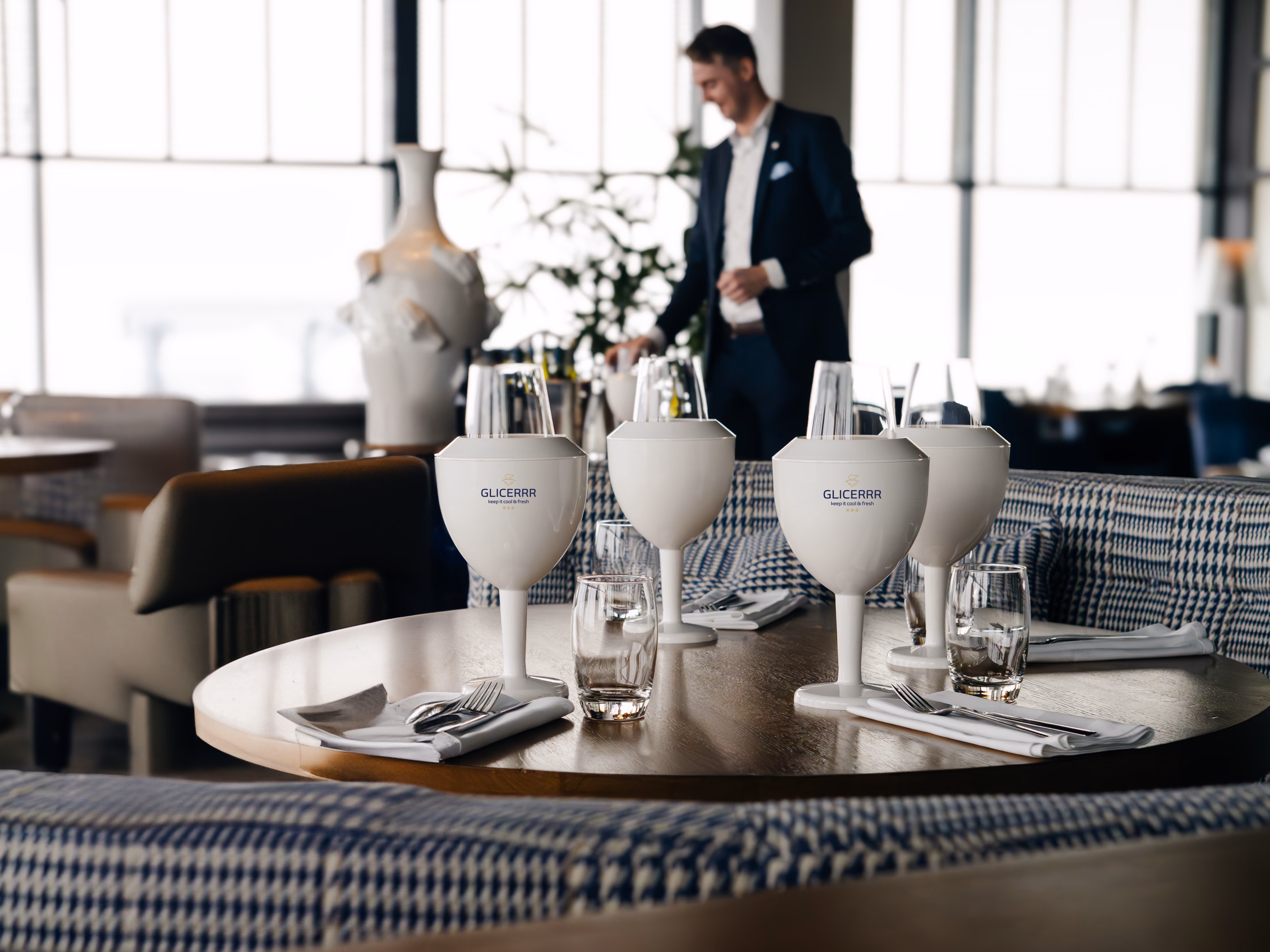 Round wooden table set for four with white Glicerrr-branded wine glasses cooler, folded napkins with cutlery, and wine glasses, in a modern restaurant with a man in a suit blurred in the background.