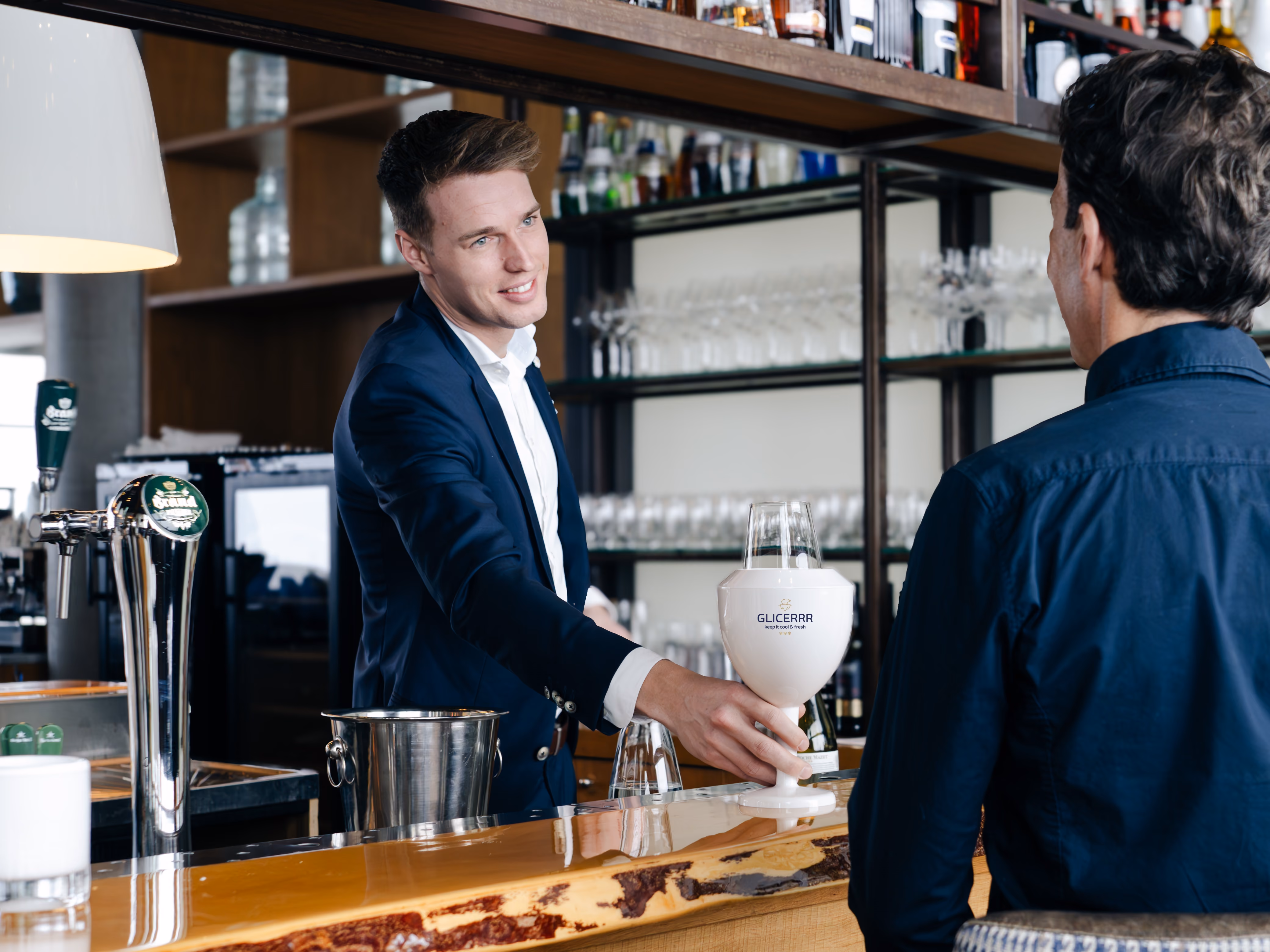 Bartender in a dark blue suit handing a GLICERRR wine glass cooler to a customer at a bar counter with shelves of bottles in the background.