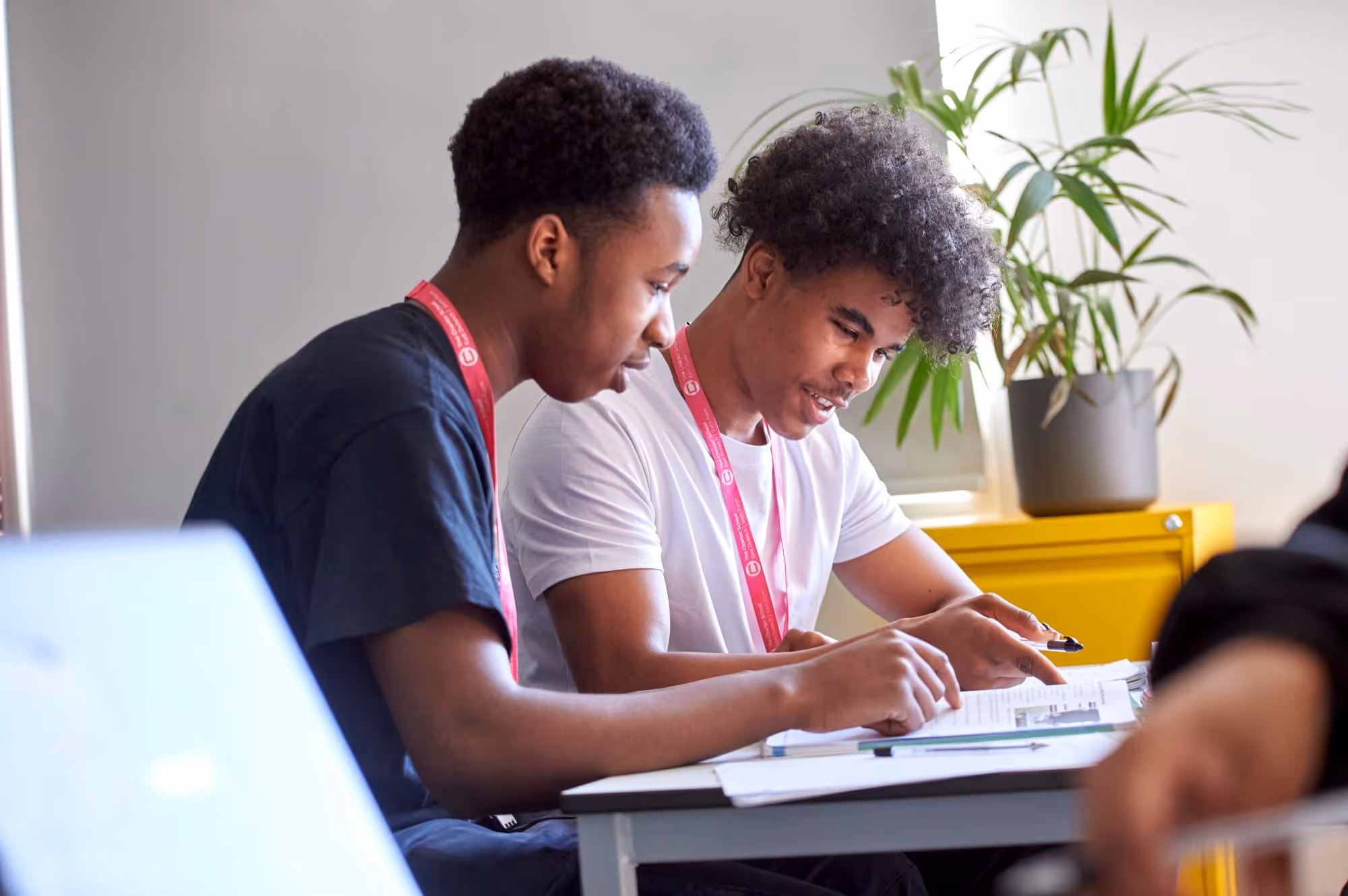 Two young men sitting at a table studying with papers and pens, one pointing at the paper.