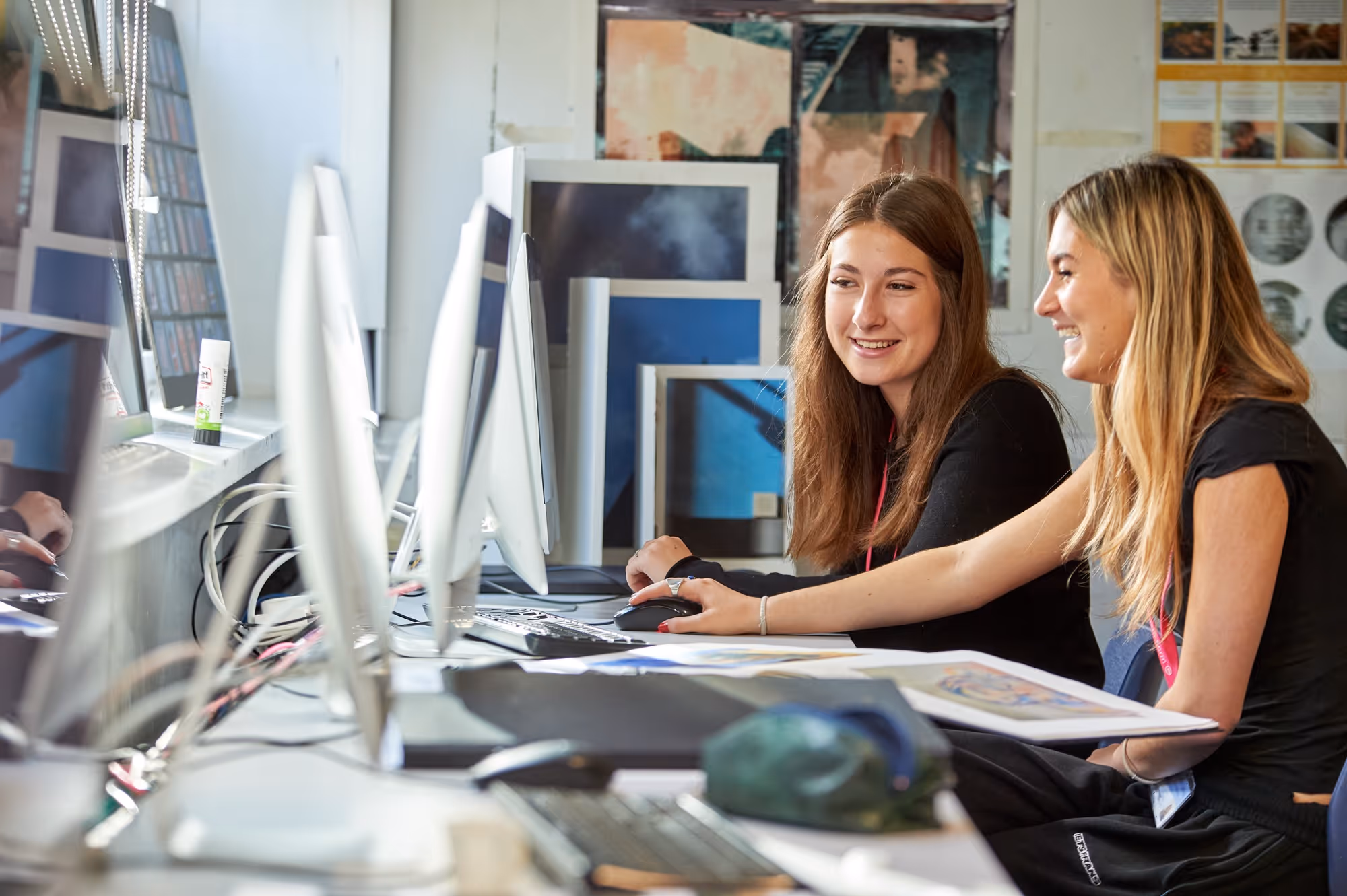 Two young women smiling and working together on a computer at a desk with papers and art prints.