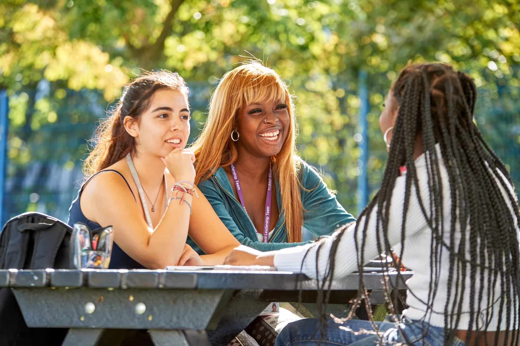 Three young women sitting outdoors at a picnic table, talking and smiling on a sunny day.