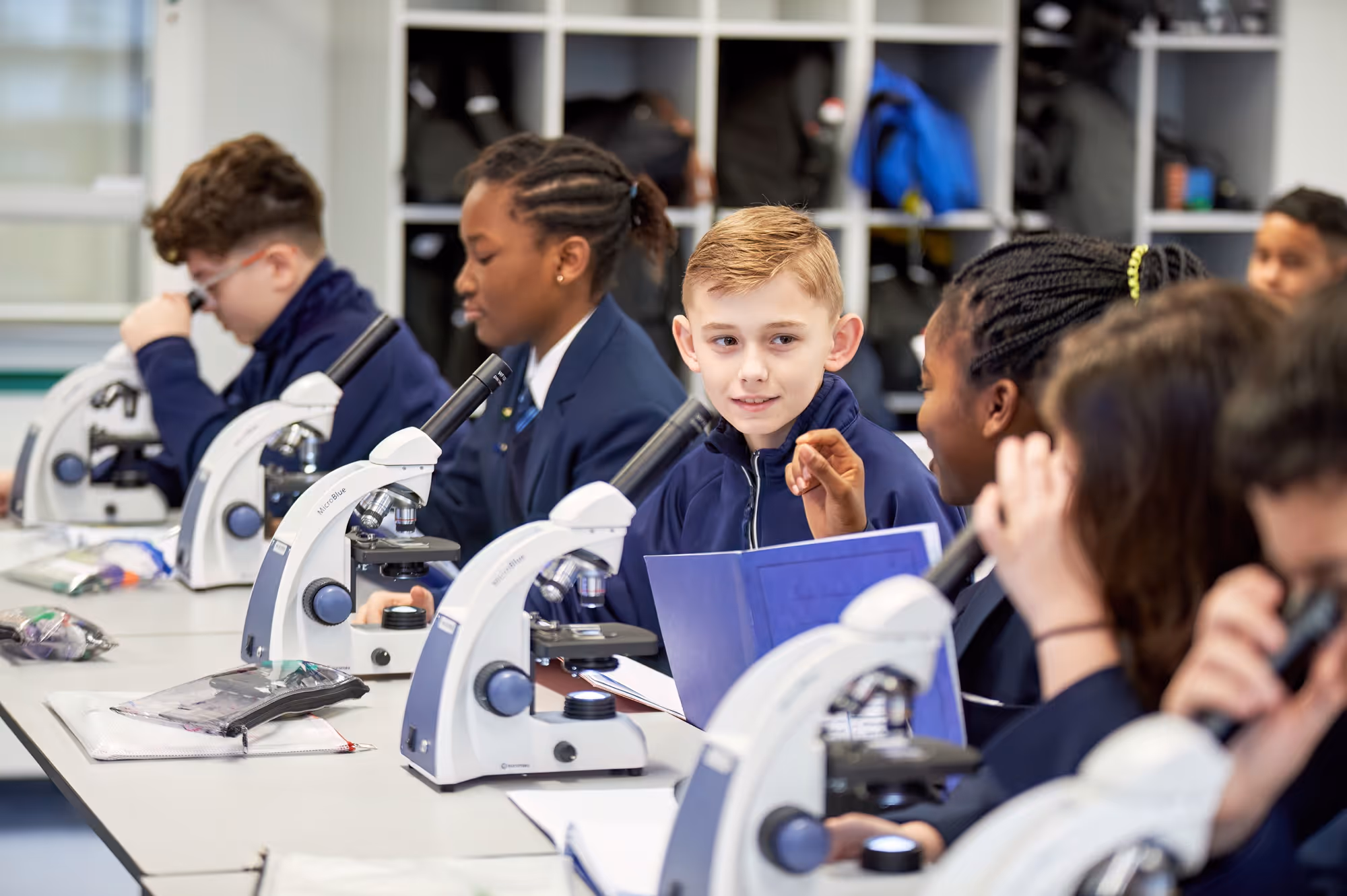 Students in school uniforms working with microscopes and discussing in a science classroom.