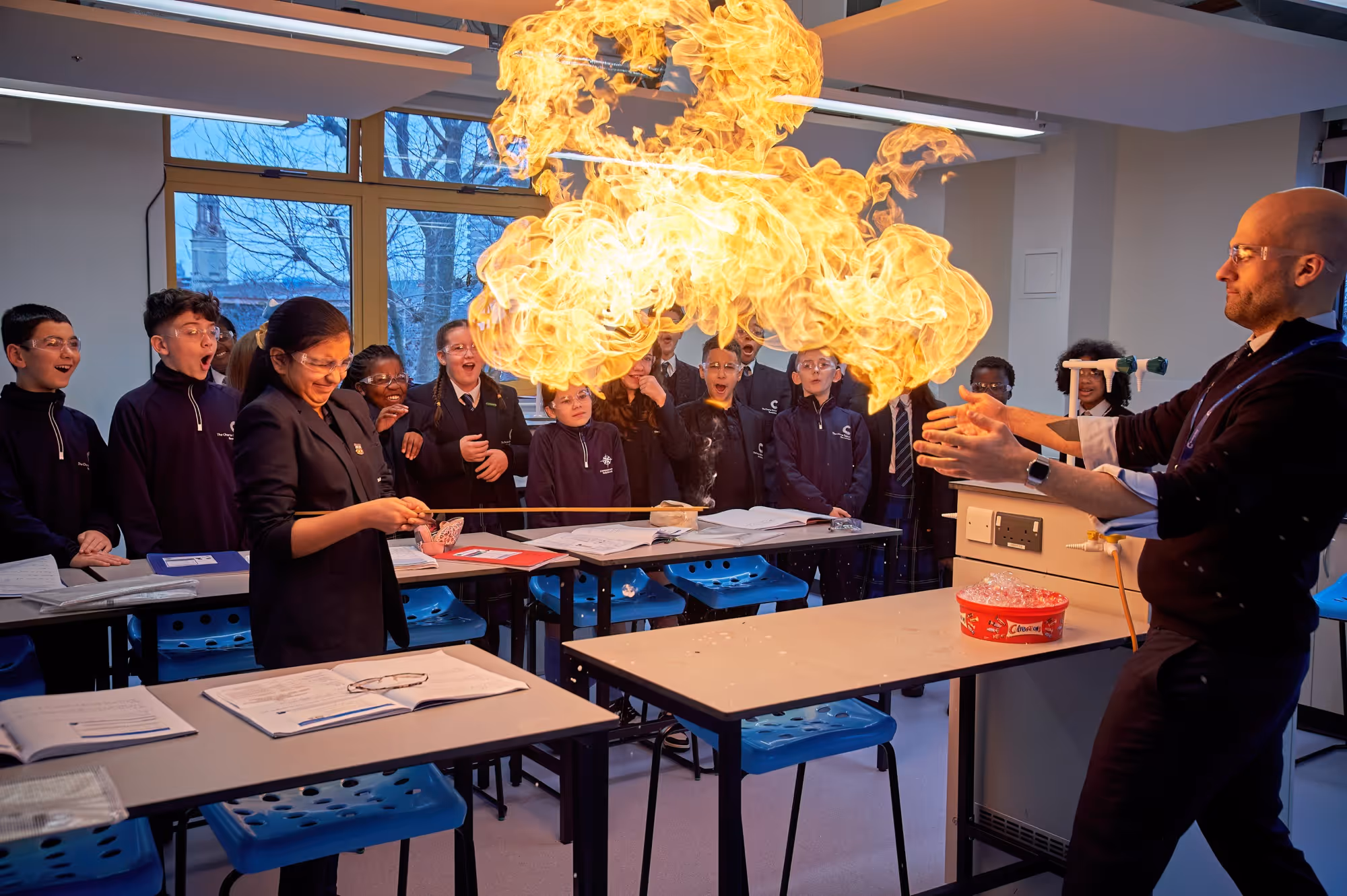 Science teacher and student creating a large fireball in a classroom experiment, classmates watching amazed.