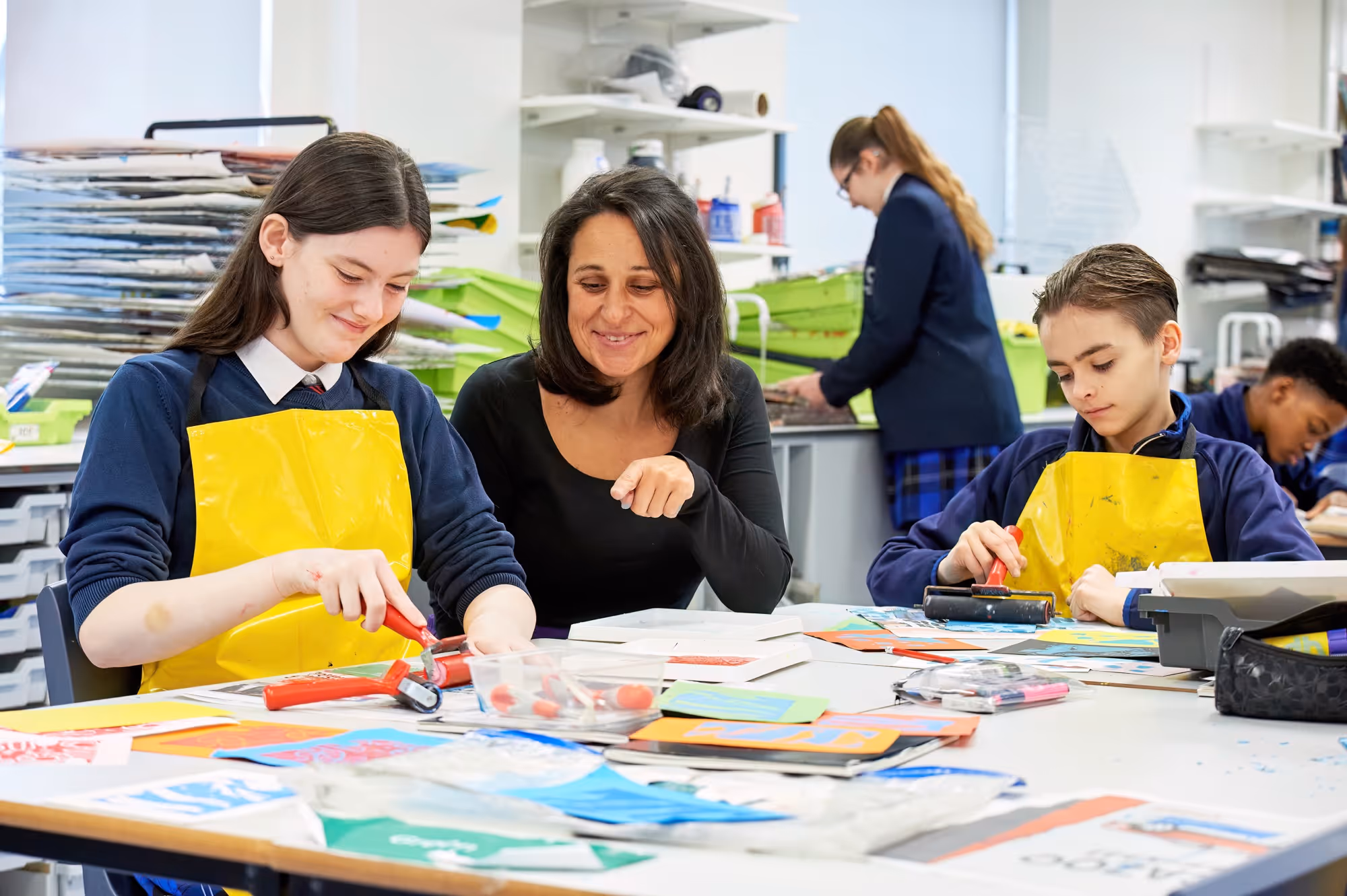 Two students in yellow aprons print art with rollers as a teacher guides them in a bright classroom.