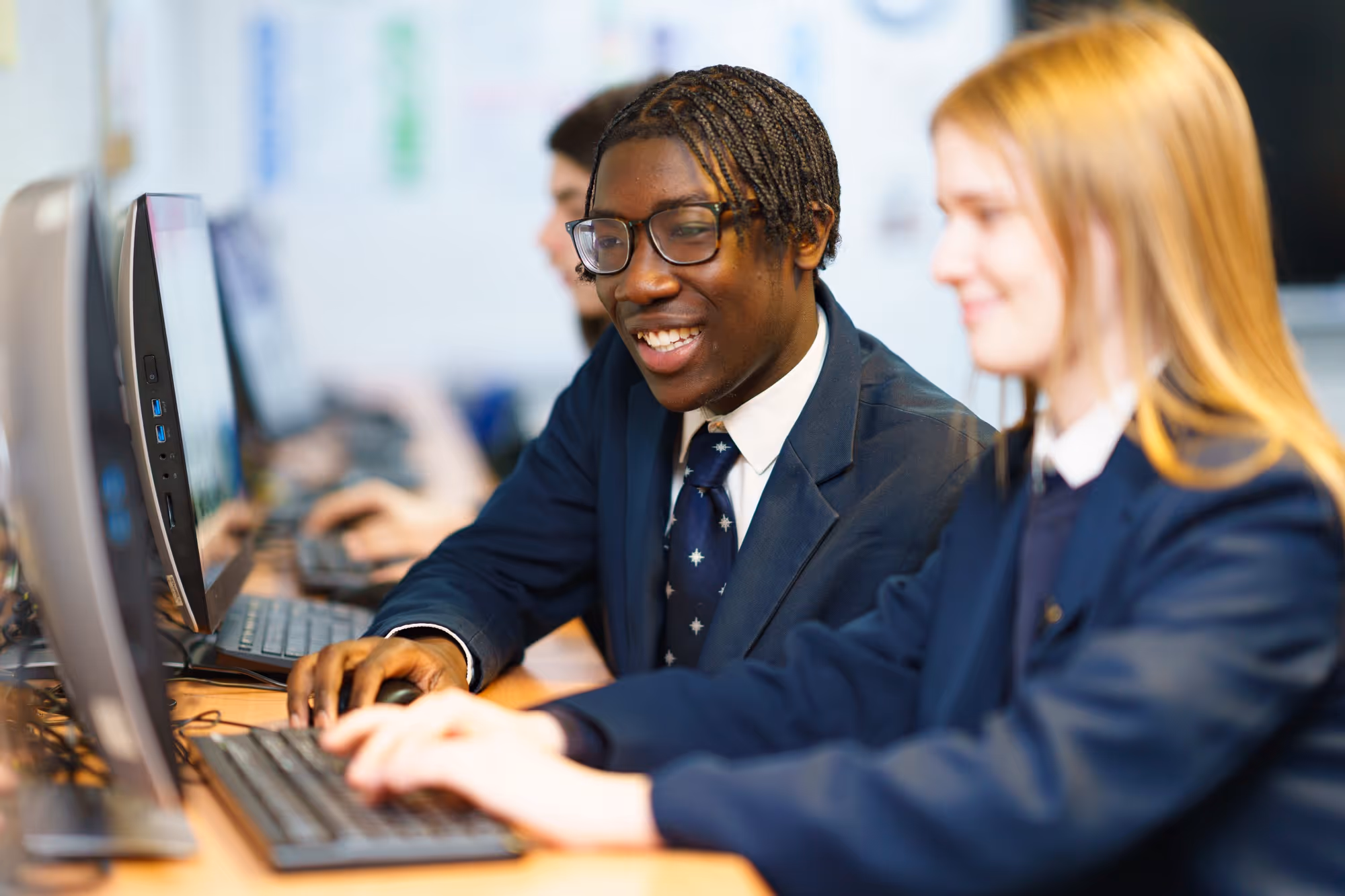 Two students in school uniforms working on desktop computers together in a classroom.