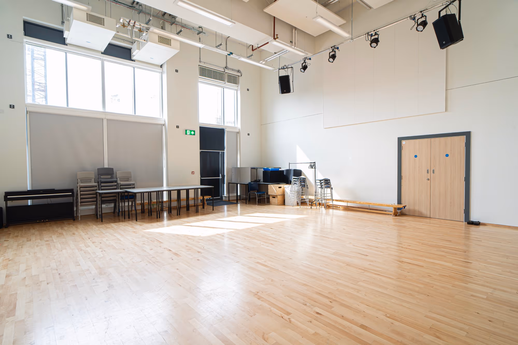 Empty bright studio room with wooden floor, stacked chairs, tables, and black piano under large windows.