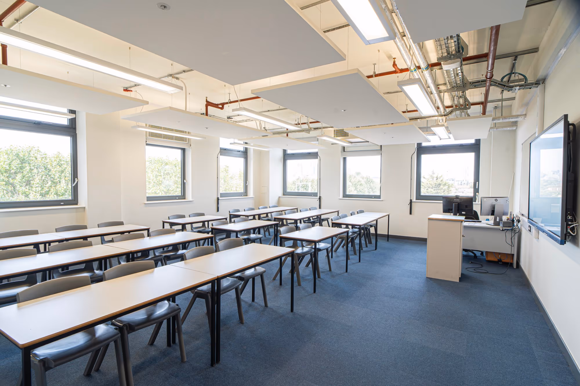 Empty modern classroom with rows of desks, gray chairs, large windows, and a teacher's desk with monitors.