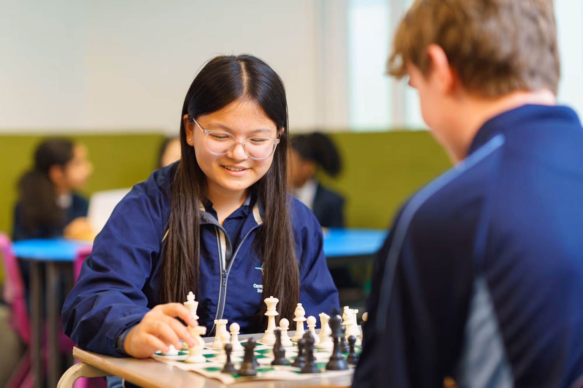 Two students playing chess in a classroom, with girl moving a white piece while smiling.