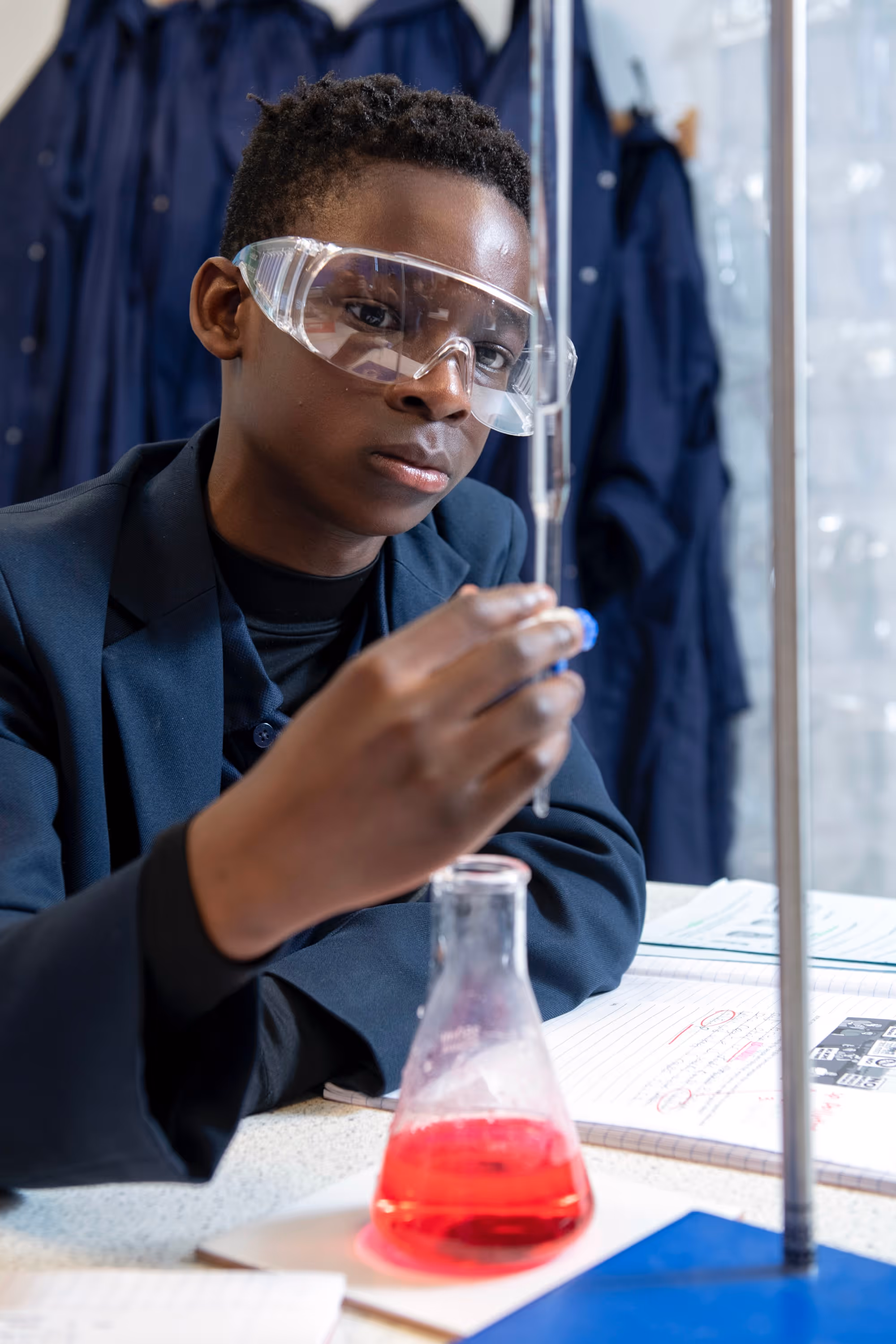 Young scientist wearing goggles holds a pipette above a flask with red liquid during an experiment.