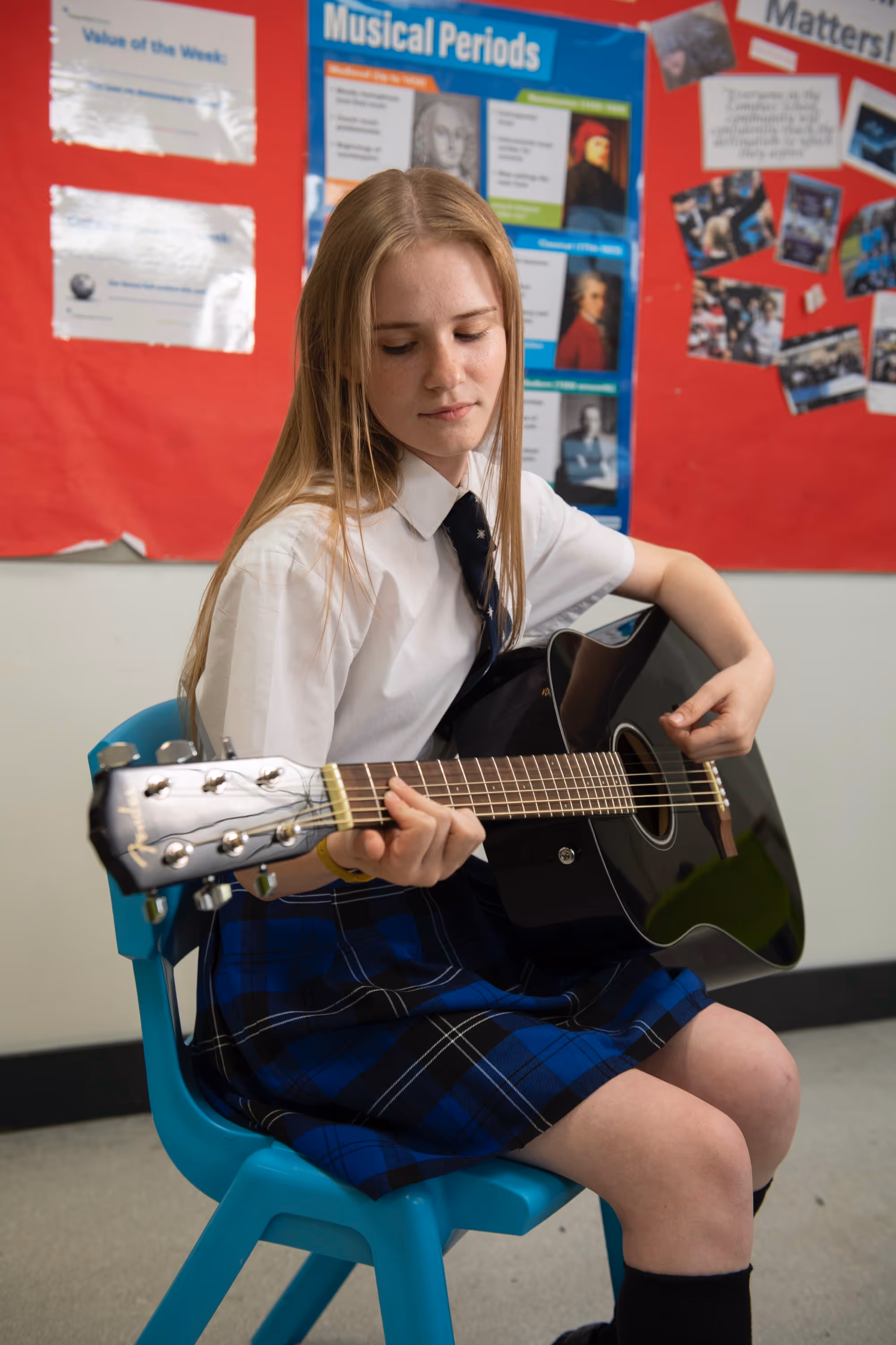 Girl in school uniform sitting on blue chair playing a black acoustic guitar in classroom.