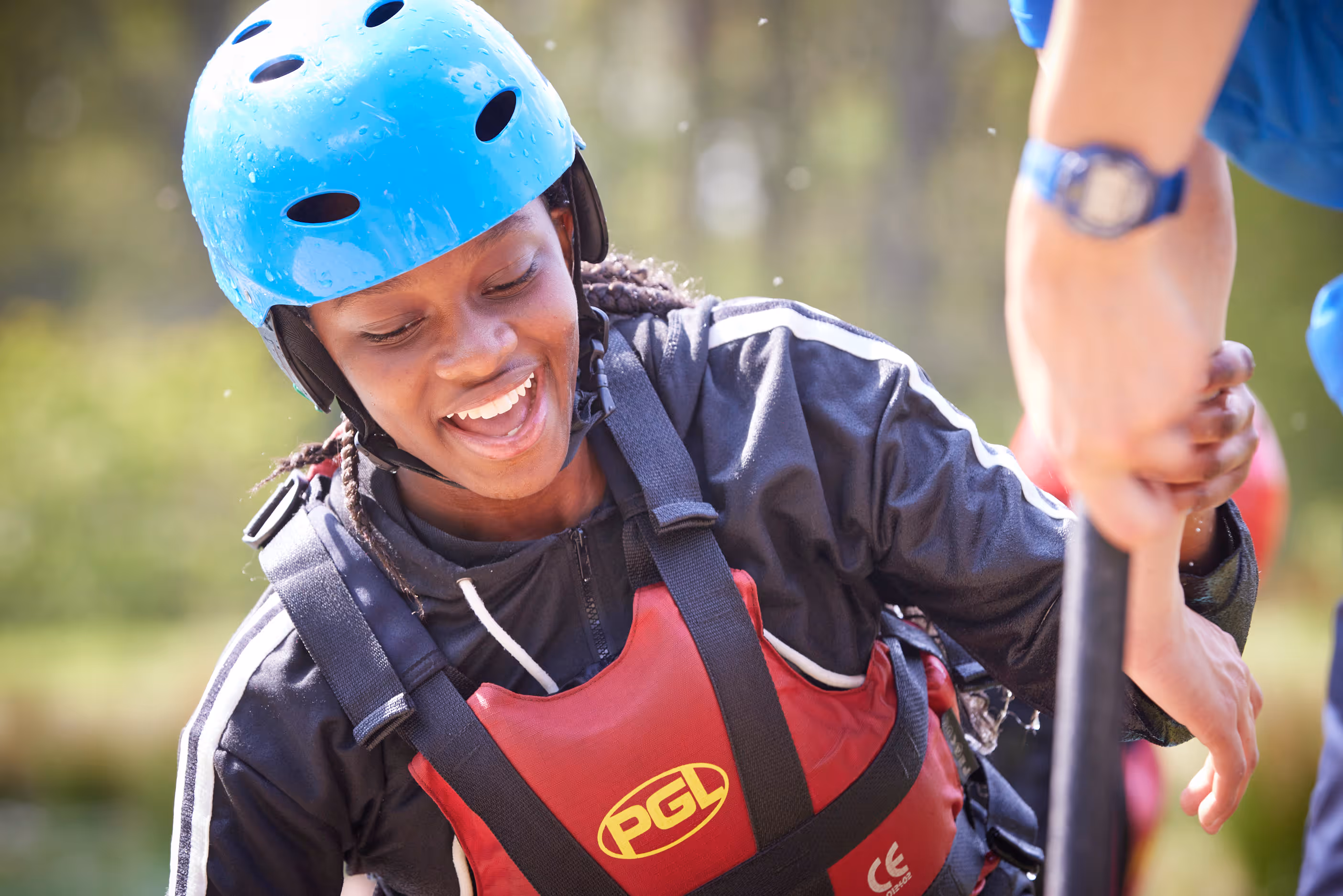 Smiling woman in blue helmet and red PGL life vest holding a paddle during outdoor water activity.