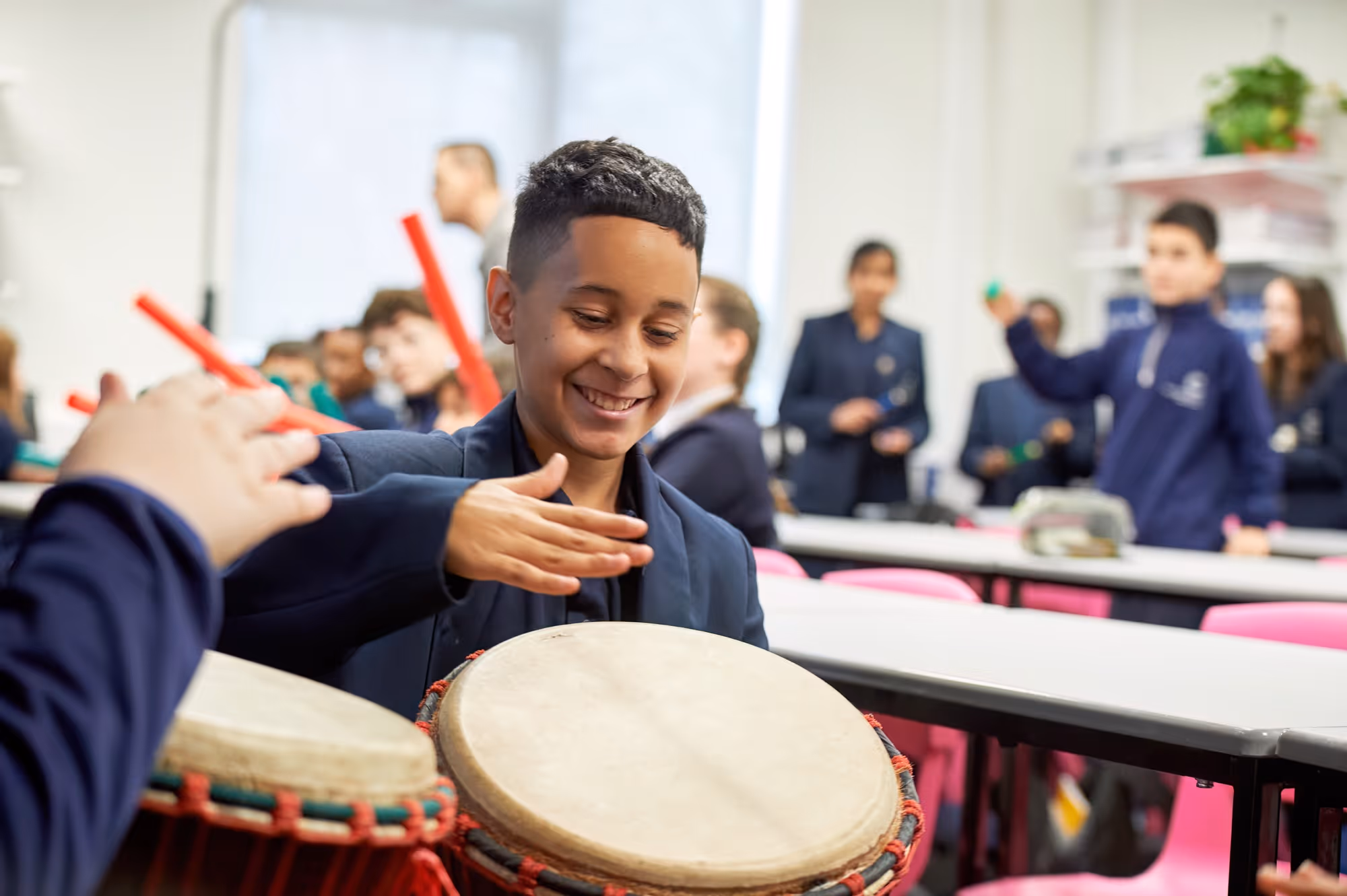 Smiling boy in a classroom playing a djembe drum with peers holding rhythm sticks in the background.