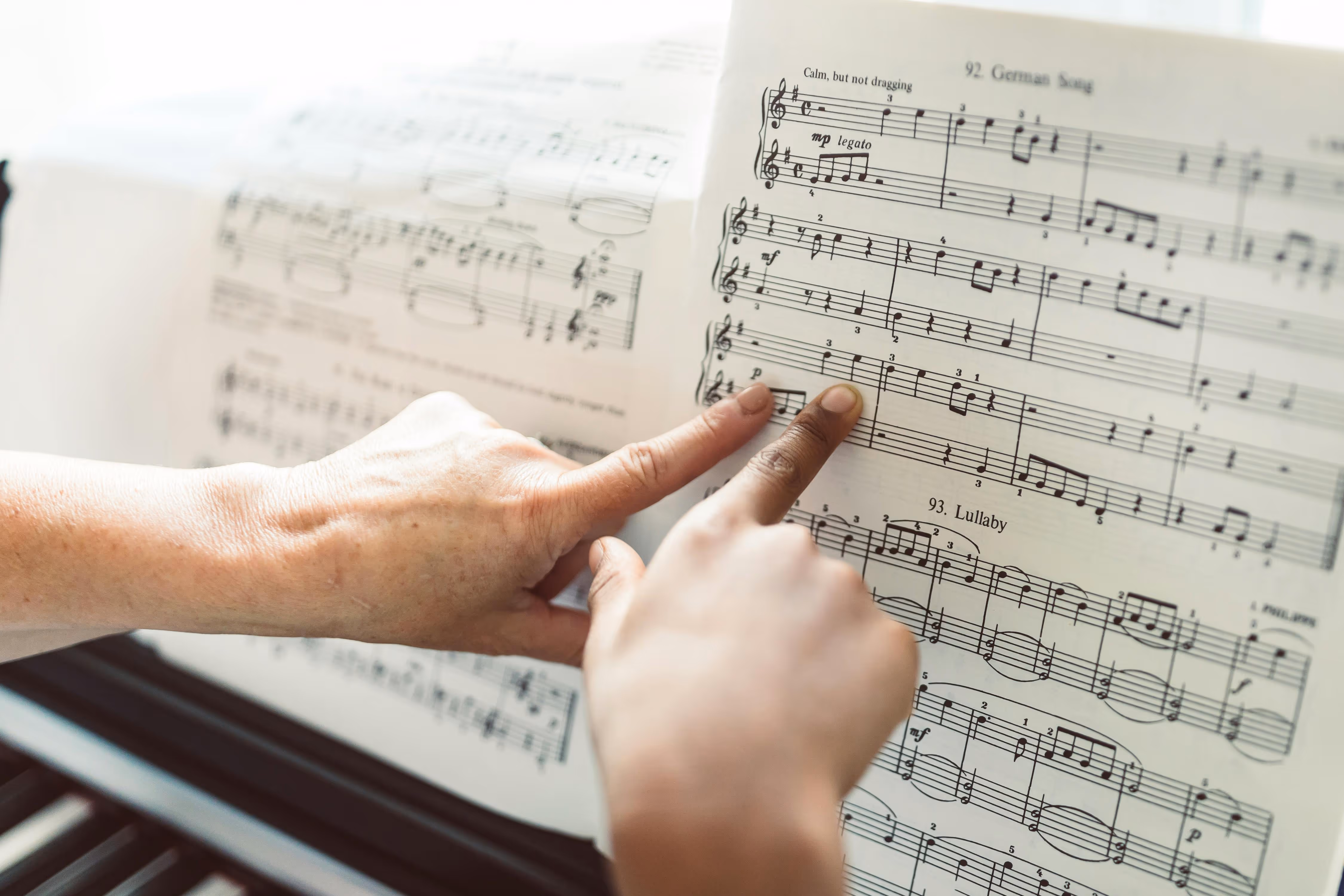 Two hands pointing at notes on sheet music titled German Song and Lullaby on a piano stand.