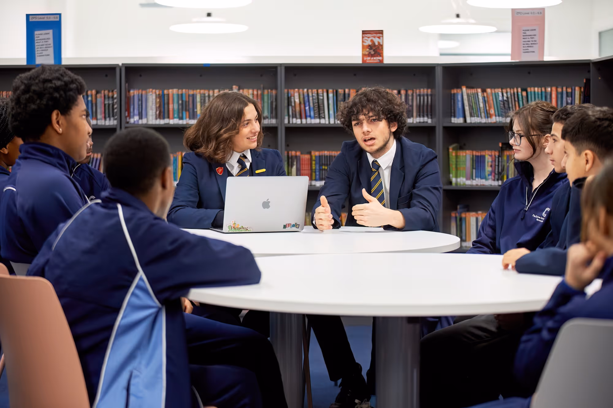 Students in school uniforms discuss around a round table in a library with bookshelves behind them.