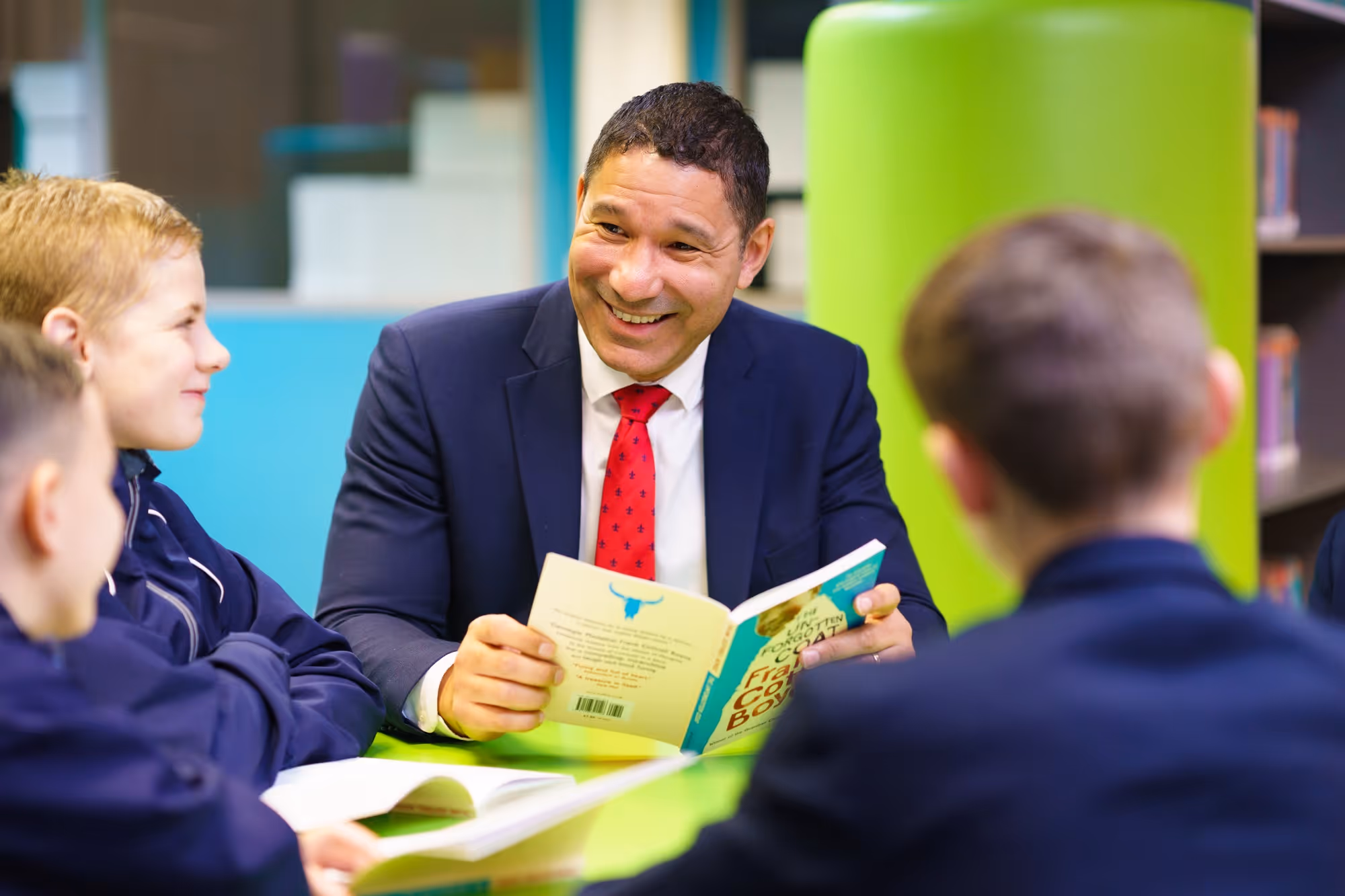 Smiling man in suit reads a book to three children in a colorful school library.