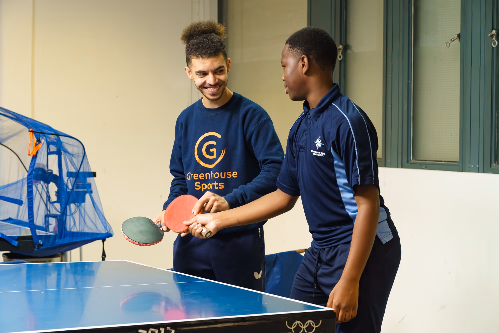 Two young men play table tennis indoors, one wearing Greenhouse Sports sweatshirt, the other compass school shirt.