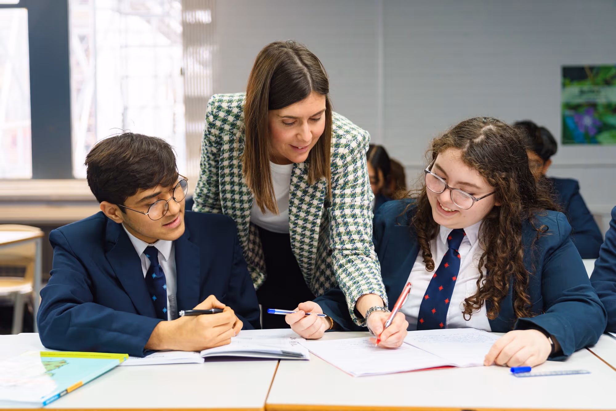 Teacher helps two students in school uniforms with their work at a classroom desk.