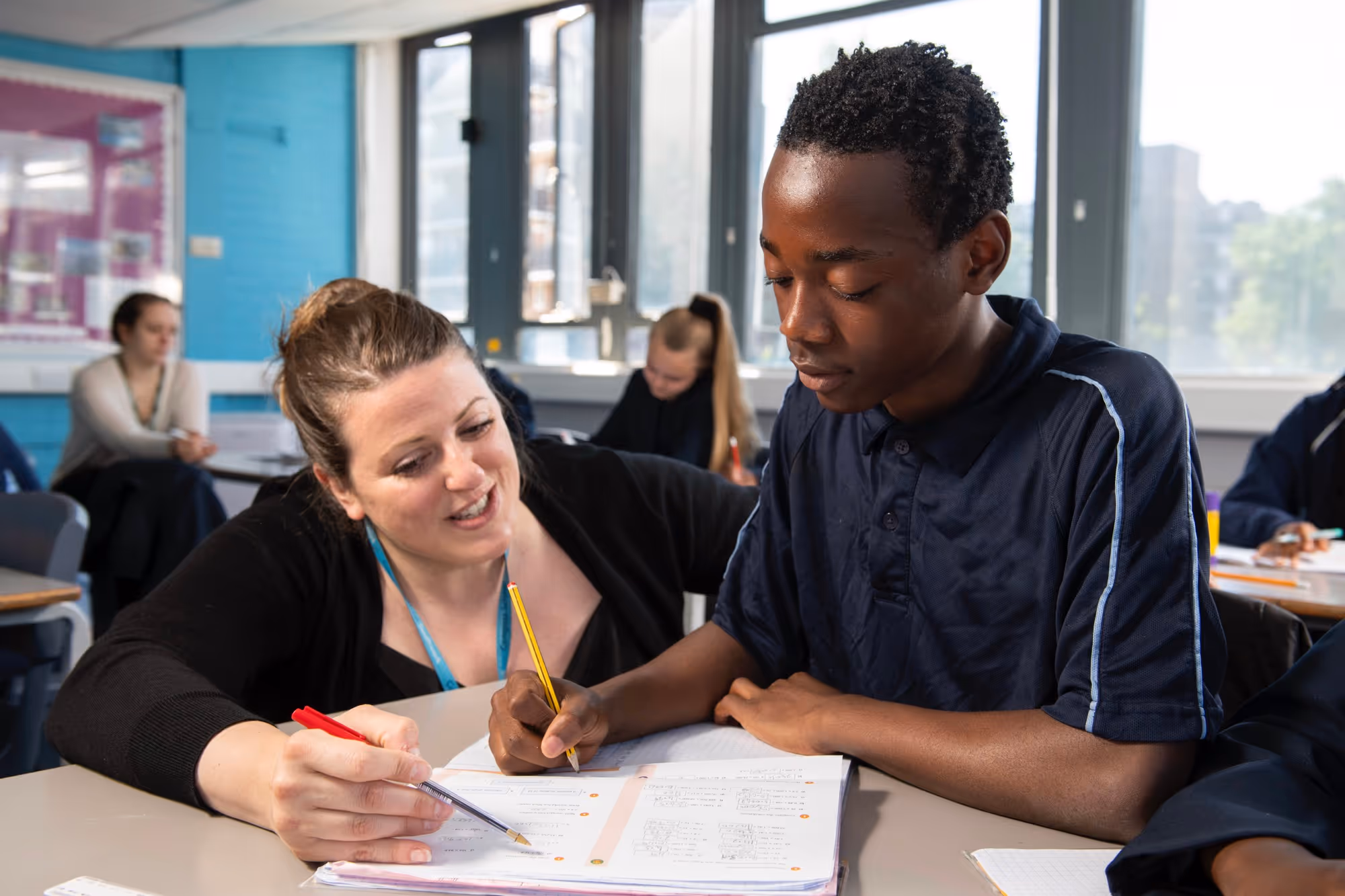 Teacher helps student with homework in classroom with other students working in background.