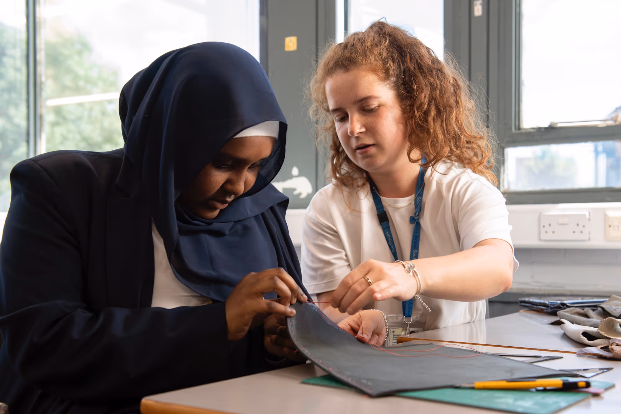 Two women working together, sewing a piece of fabric at a table indoors near windows.