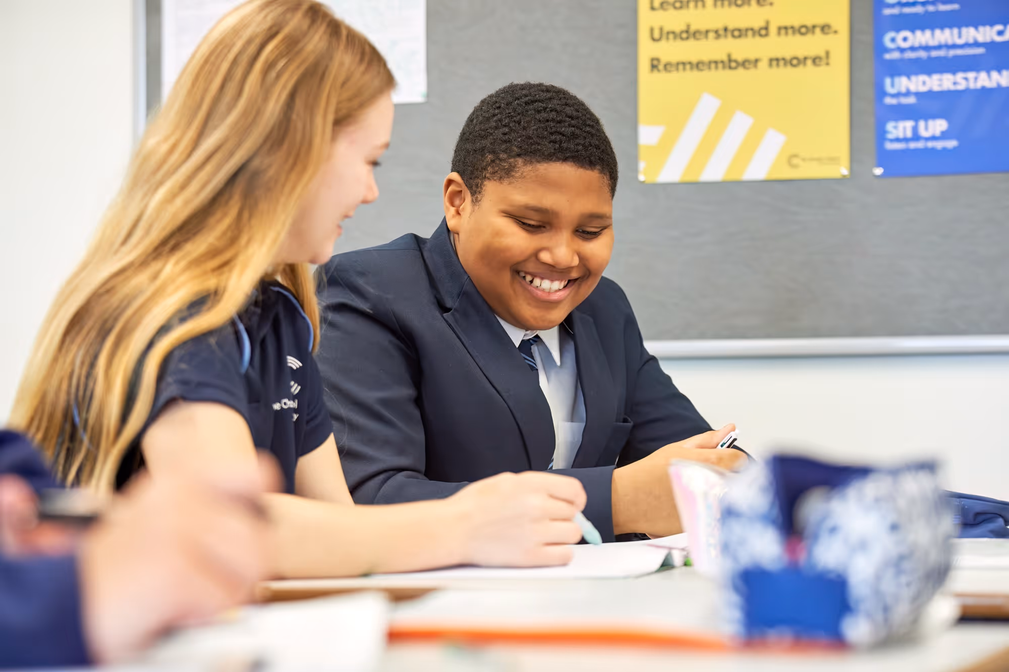Smiling boy in school uniform with a girl in a classroom, working on a notebook together.