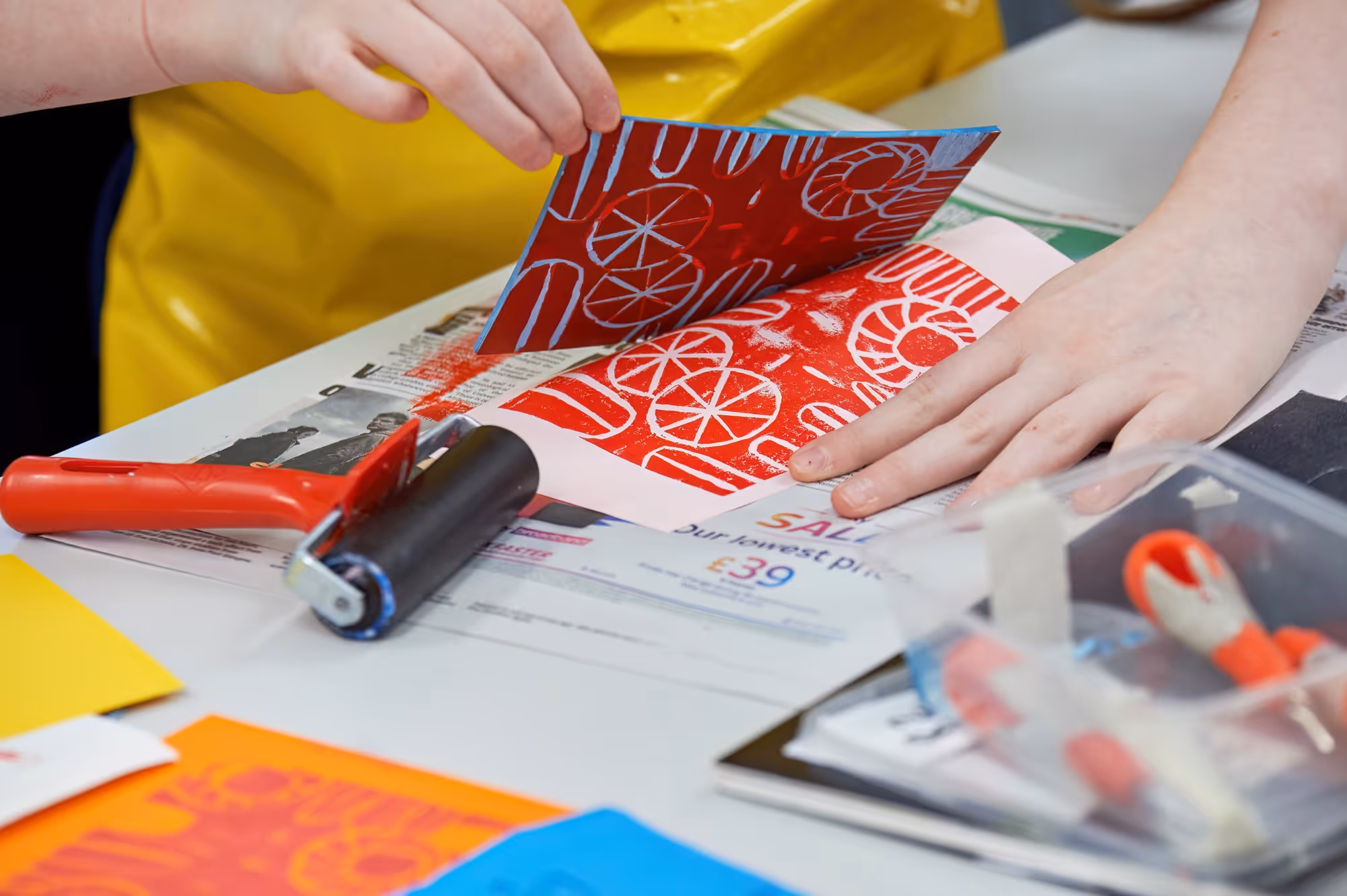 Hands lifting a red ink print from paper with white circular and oval designs, next to a brayer roller.