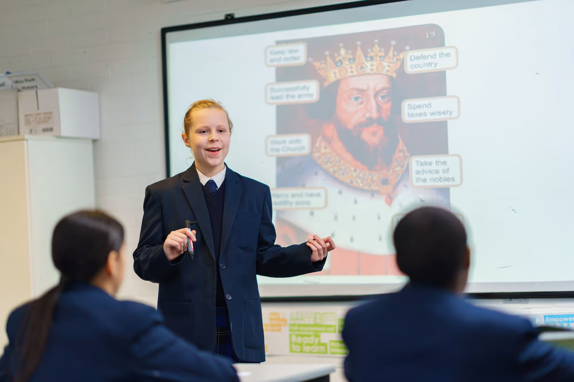 A student in uniform giving a presentation in class with a historic king image on the screen behind her.